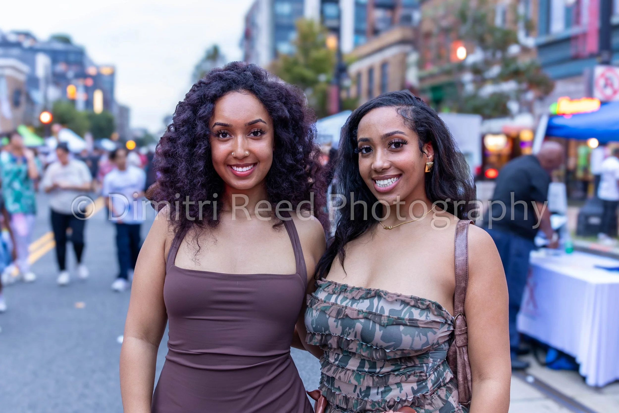 Two smiling women with dark hair and tan skin standing close together at an outdoor street fair or festival, with blurred people and colorful booths in the background.