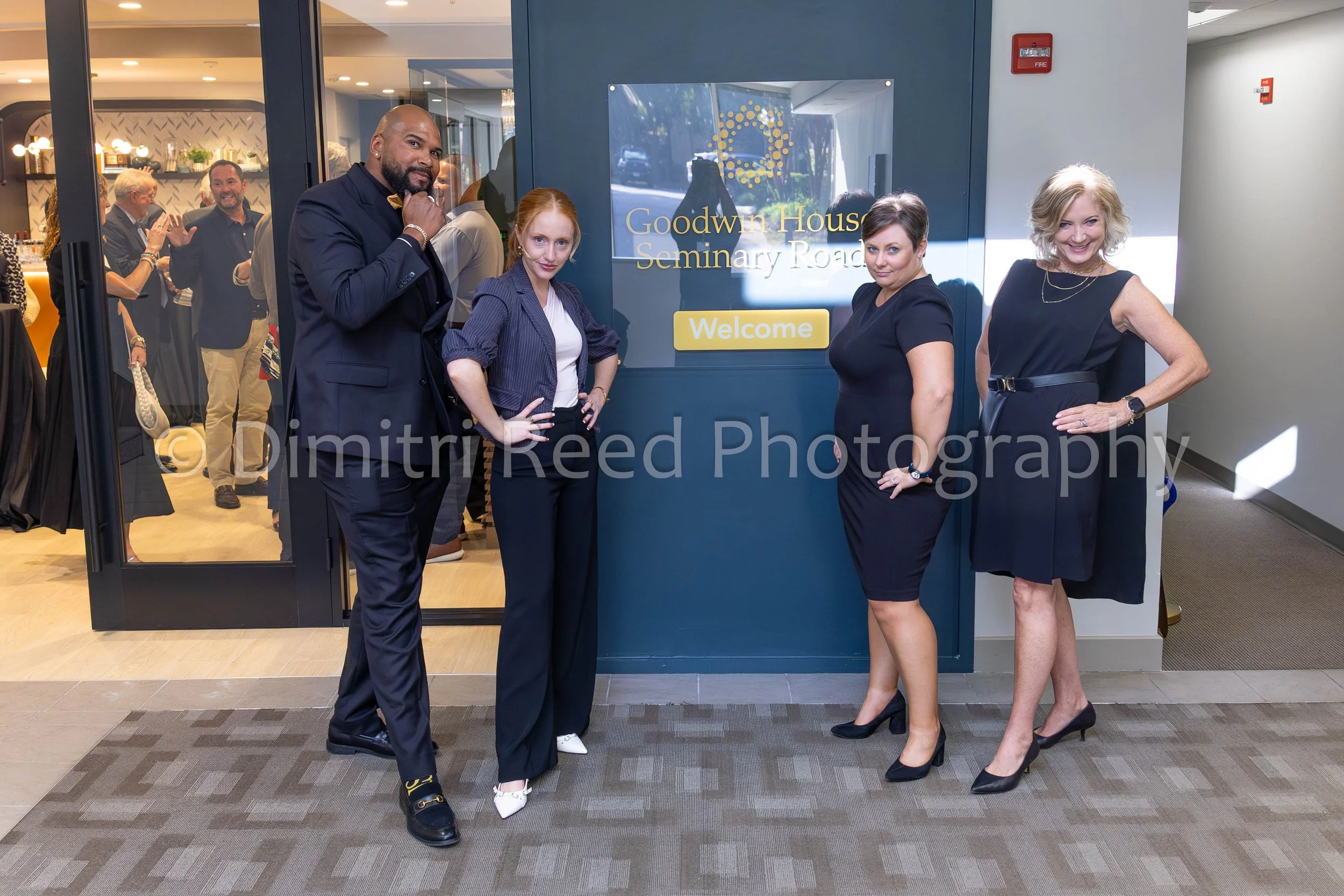 Group of four people, three women and one man, standing in front of a sign that reads 'Goodwin House Seminary Road' with a welcome message, inside a building. Other people are visible inside through the glass door.