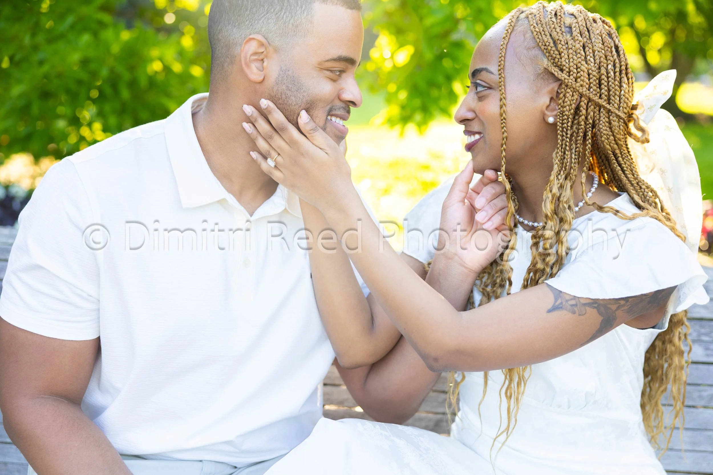 A man and woman sitting close together outdoors, smiling and touching each other's faces, with the woman gently holding the man's chin. The woman has braided hair and is wearing jewelry, and both are wearing white clothing, surrounded by green foliag