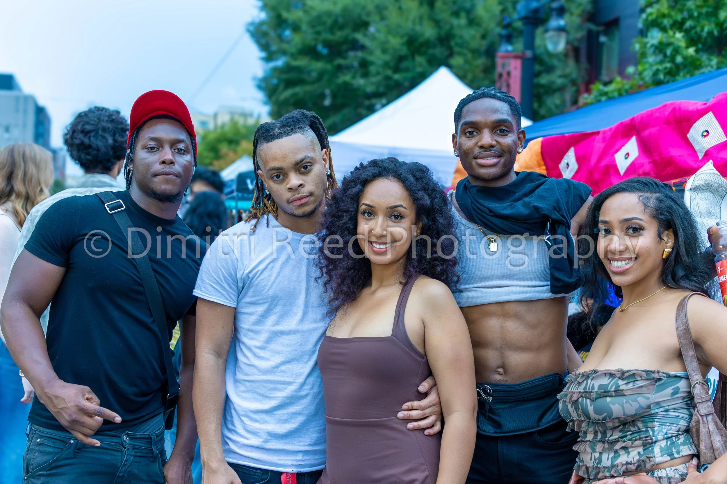 Group of young people posing at an outdoor event, with tents and trees in the background, smiling and looking at the camera.