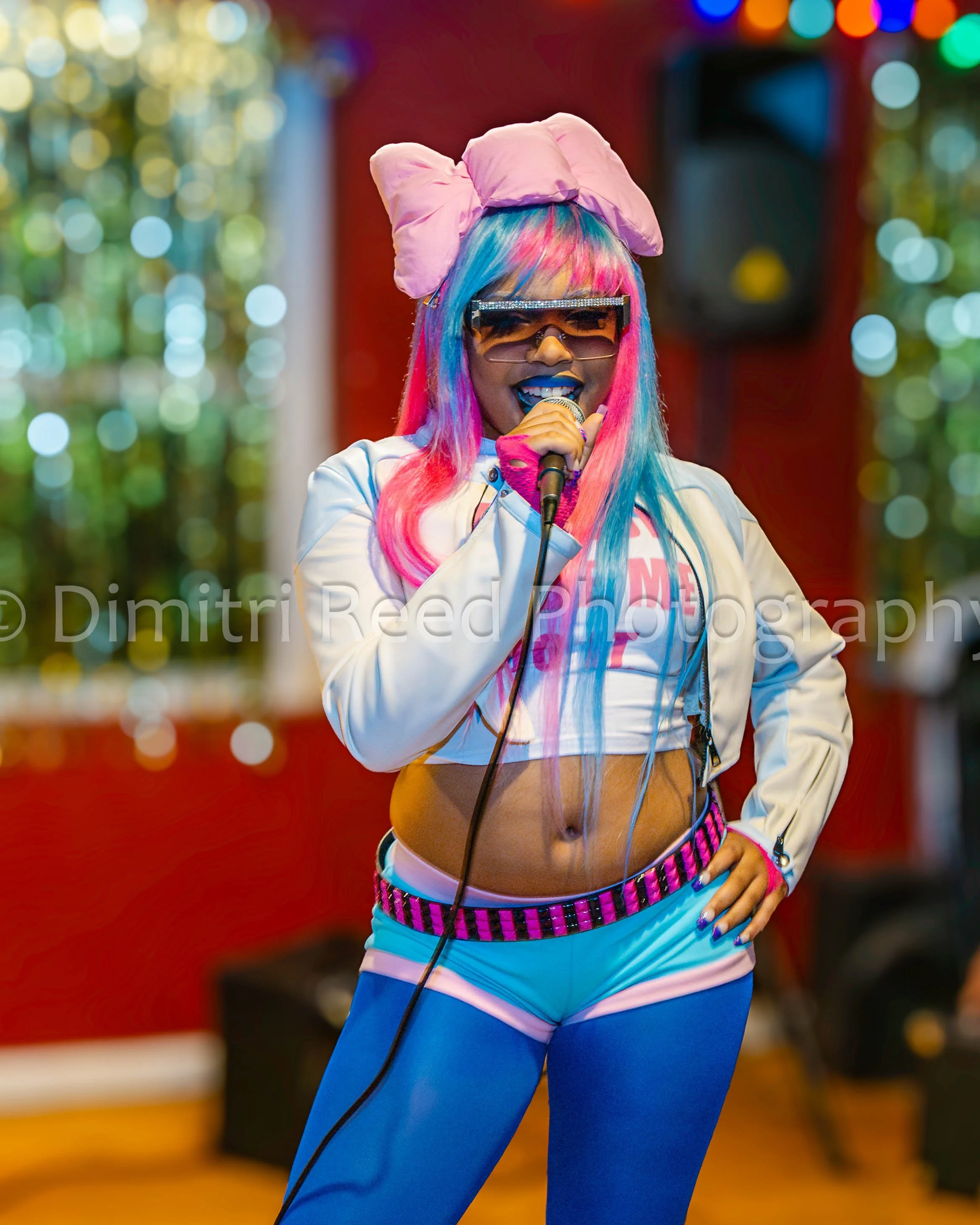 A woman with brightly colored pink, blue, and yellow hair, wearing large sunglasses with a sparkly frame, and an oversized pink bow headband. She is holding a microphone, dressed in a white athletic jacket with colorful trim, and bright blue pants. S