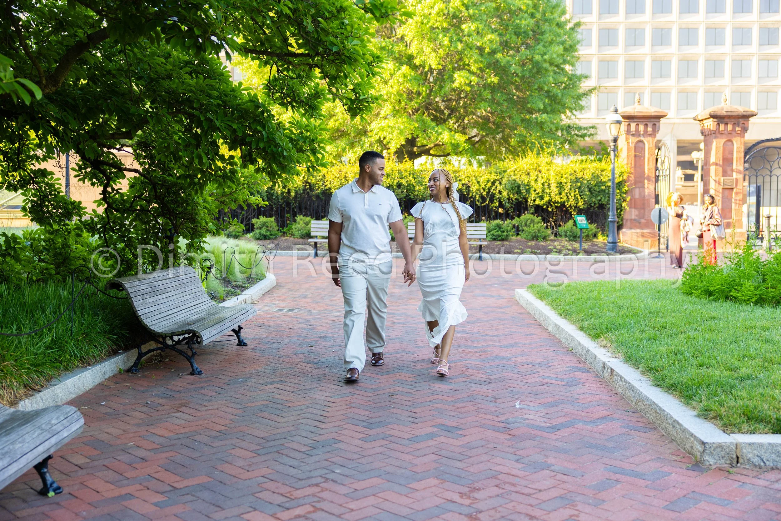 A happy couple with dark skin walking hand in hand through a city park on a sunny day. The man is dressed in a white shirt and pants, and the woman wears a white dress. They are smiling at each other as they walk along a brick pathway, surrounded by 
