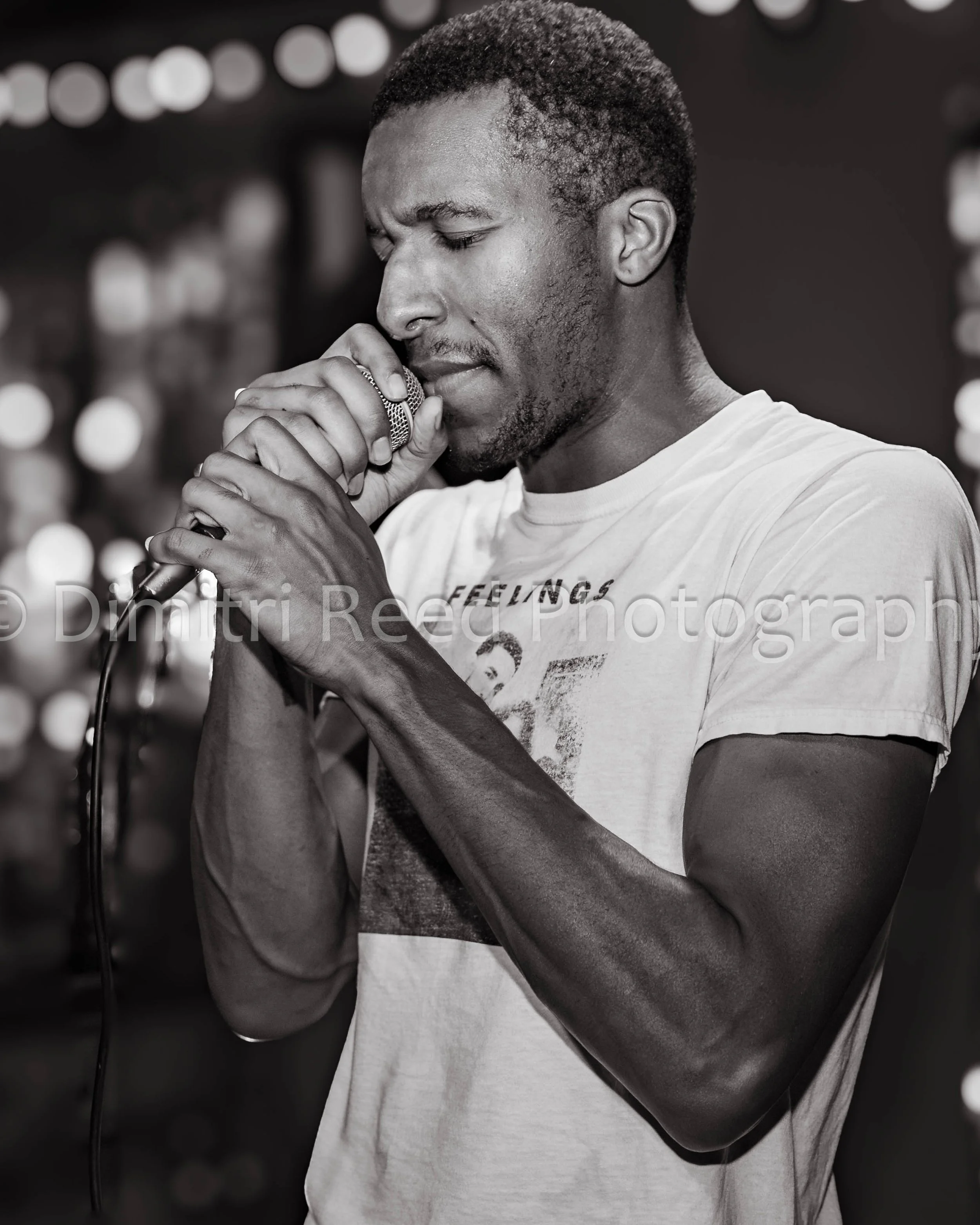 A young man with short curly hair and a beard, holding a microphone with both hands, eyes closed, appearing to sing or speak passionately.