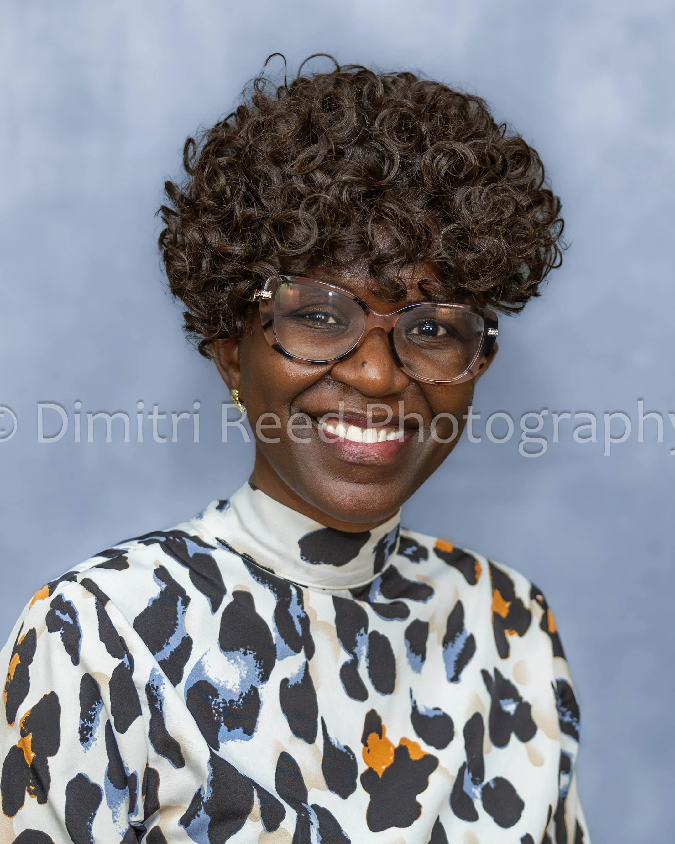 Close-up of an African American woman with short, curly hair, wearing glasses and a patterned top, smiling against a plain, light blue background.