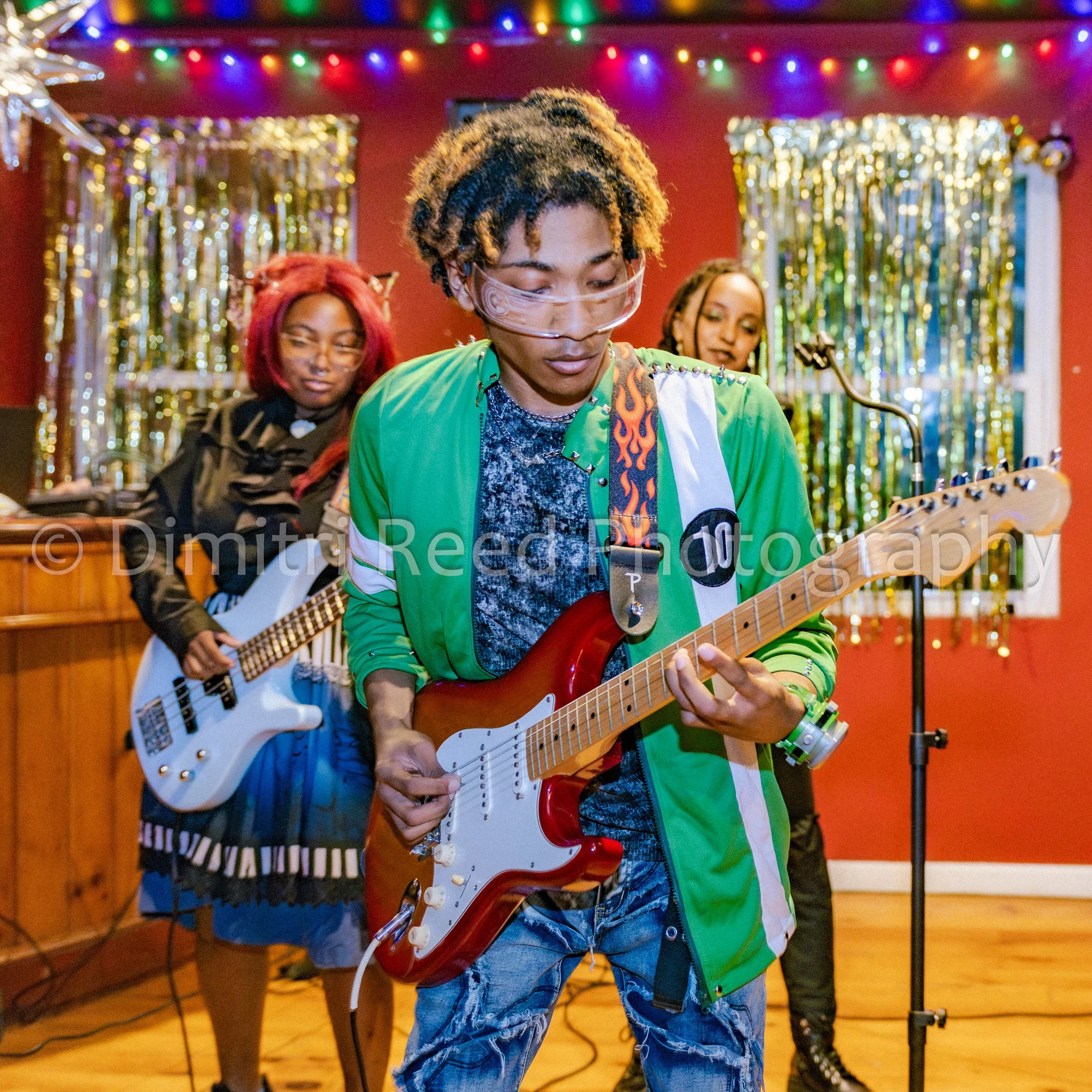 Three musicians performing at a festive event, with colorful decorative lights and gold tinsel in the background. The man in the center plays an electric guitar, wears a green jacket, and has blonde and black dreadlocks. Two women, one with red hair 