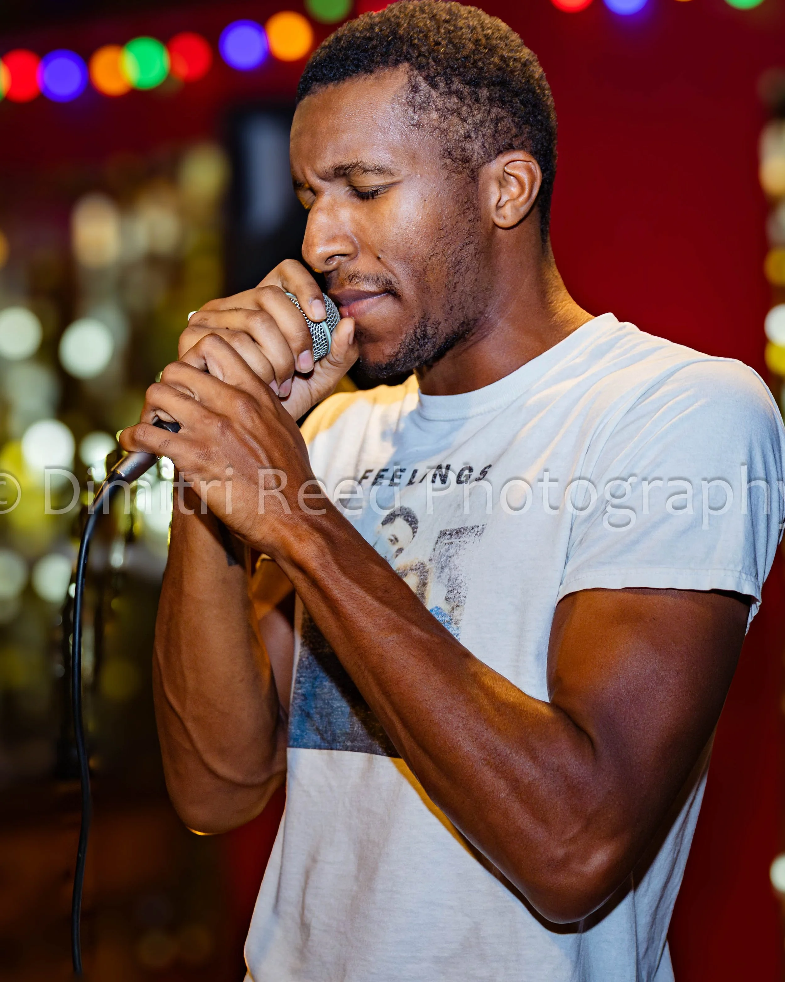 A young man with short curly hair and a light beard is singing into a microphone with his eyes closed, wearing a white T-shirt with a graphic print, in a colorful, blurred background of lights.