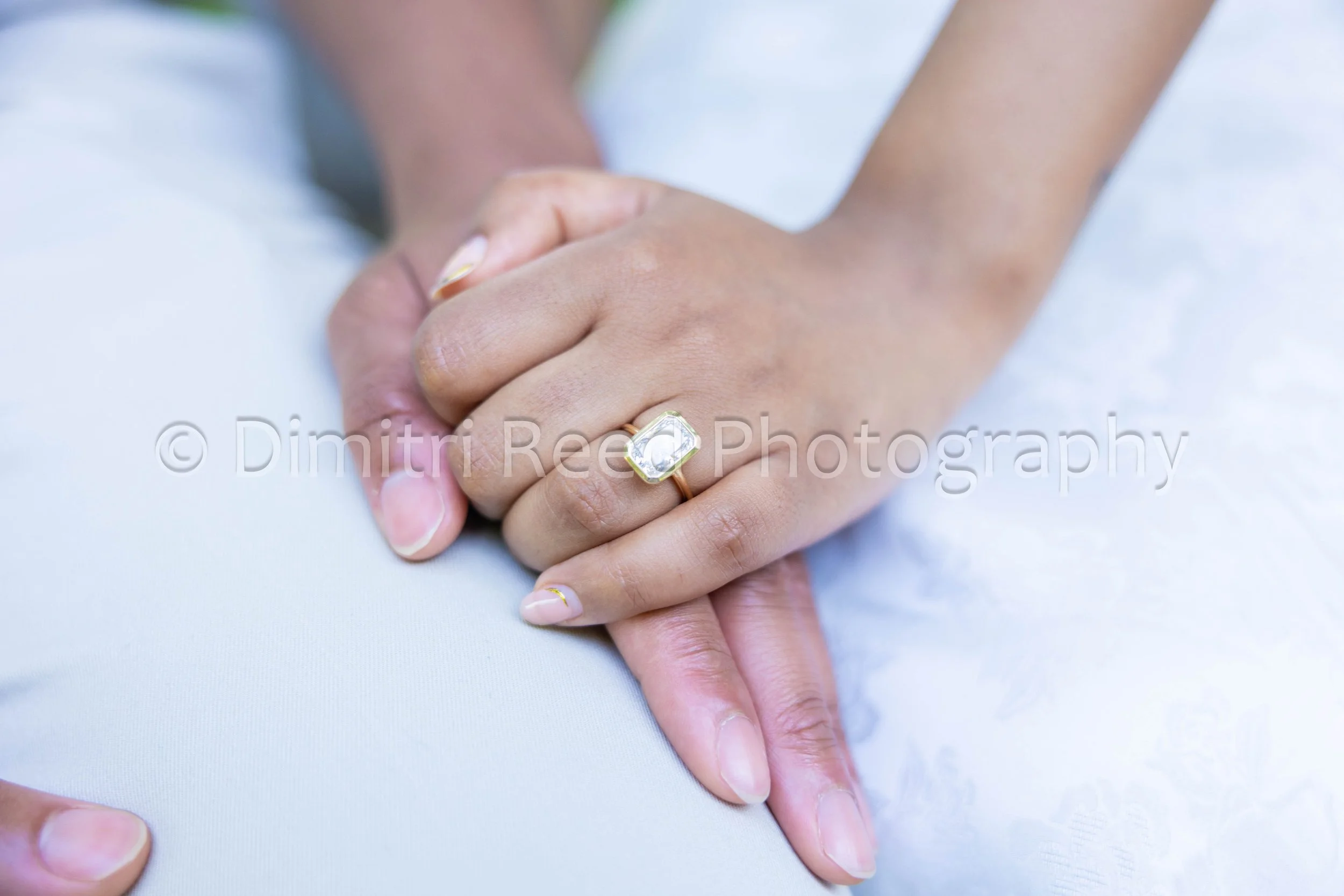 Close-up of two hands holding each other, with one hand wearing a large clear gemstone ring and the other hand showing a gold band. The background is blurred, and the focus is on the hands and rings.