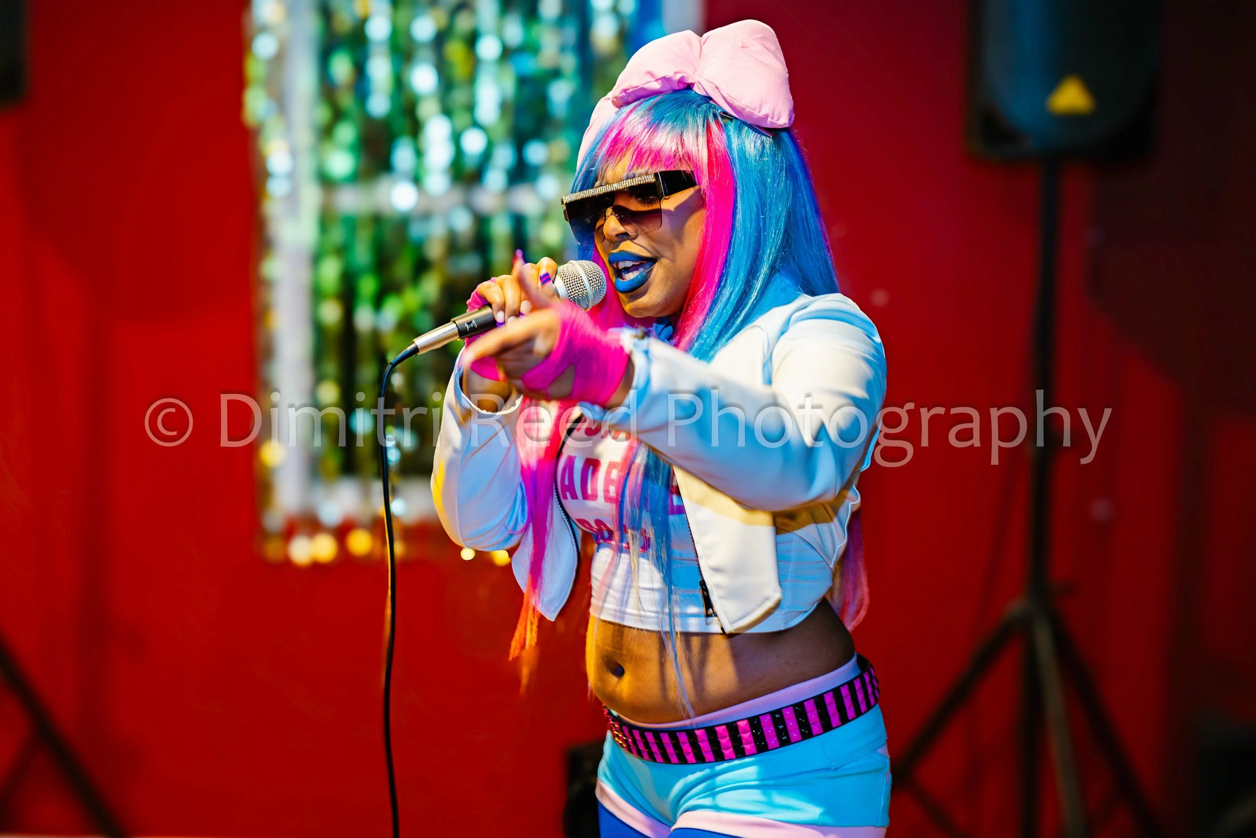 A woman with colorful hair, wearing sunglasses, a pink hat, a white jacket, and shorts, singing into a microphone indoors.