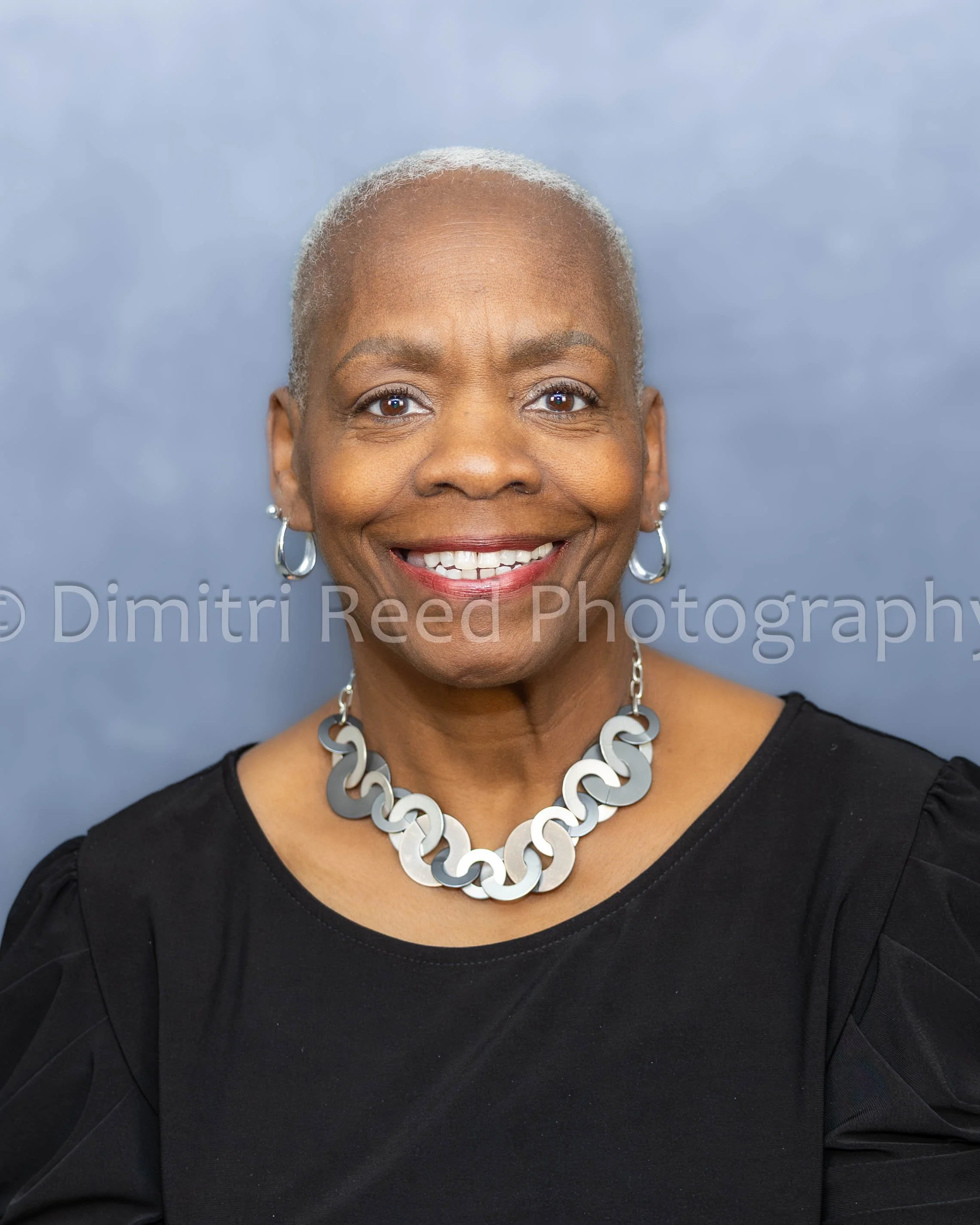 Close-up of a smiling woman with short gray hair wearing earrings and a necklace, against a blue sky background.