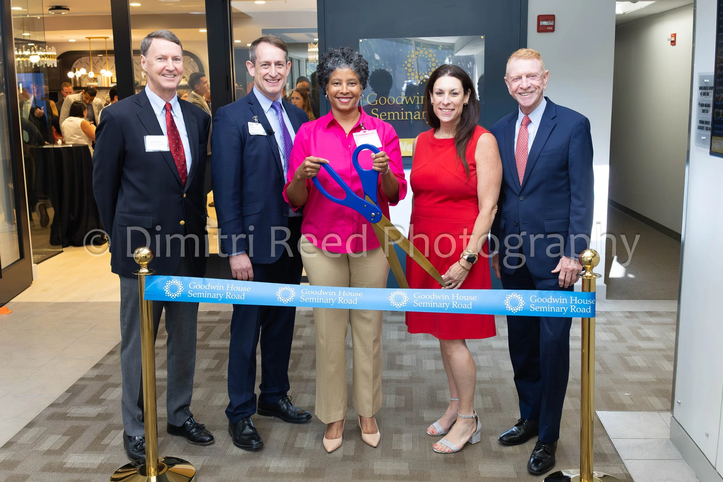 Group of five professionally dressed people, three men and two women, standing behind a ribbon with the words "Goodwin House Seminary Road," during a ribbon-cutting ceremony at an indoor event. One woman in the middle is holding a large pair of sciss