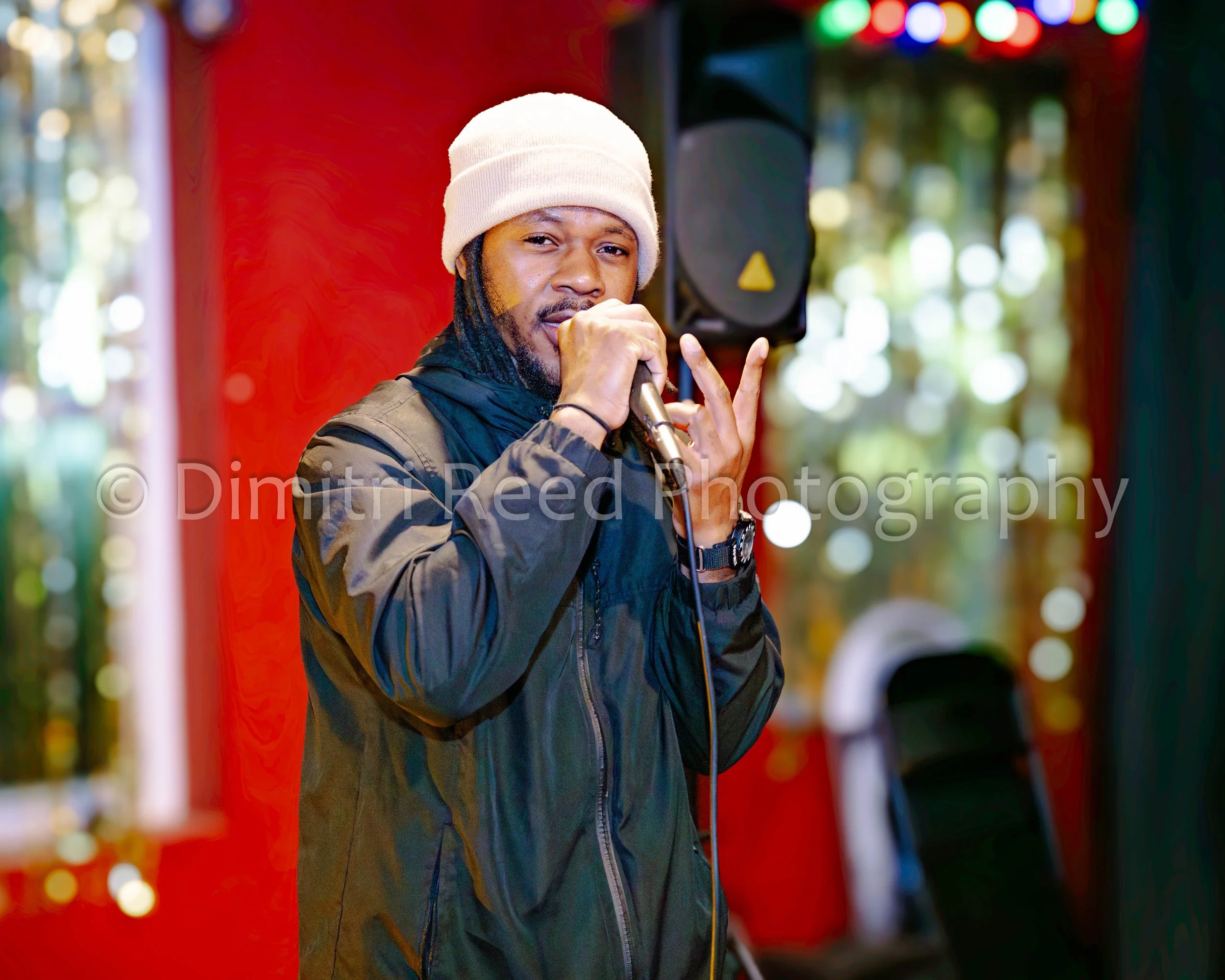 A man wearing a beige knit cap and black jacket singing into a microphone at a festive event, with colorful holiday lights in the background.