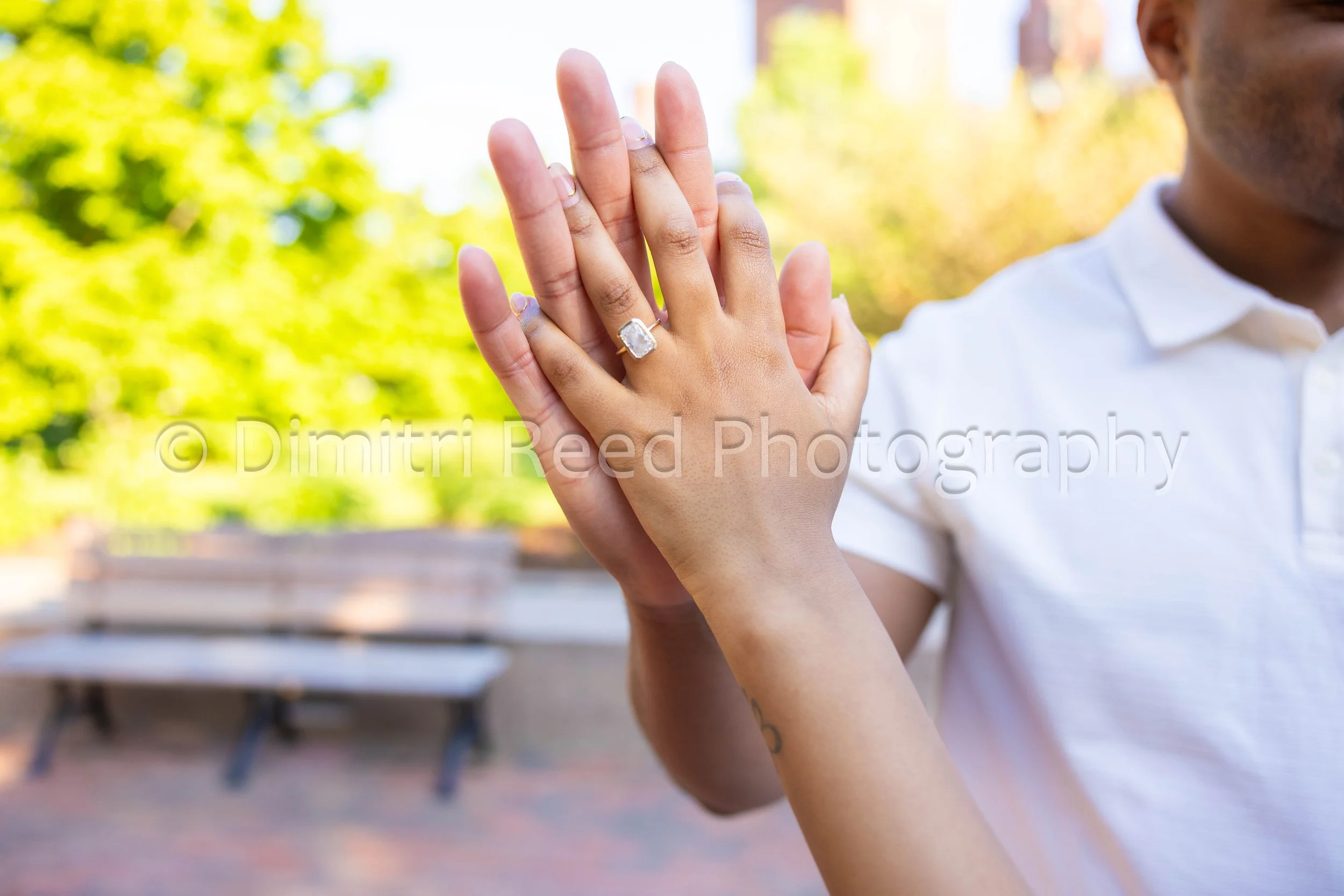 A close-up of two people, one male and one female, holding hands outdoors on a sunny day with blurred green and yellow trees in the background.