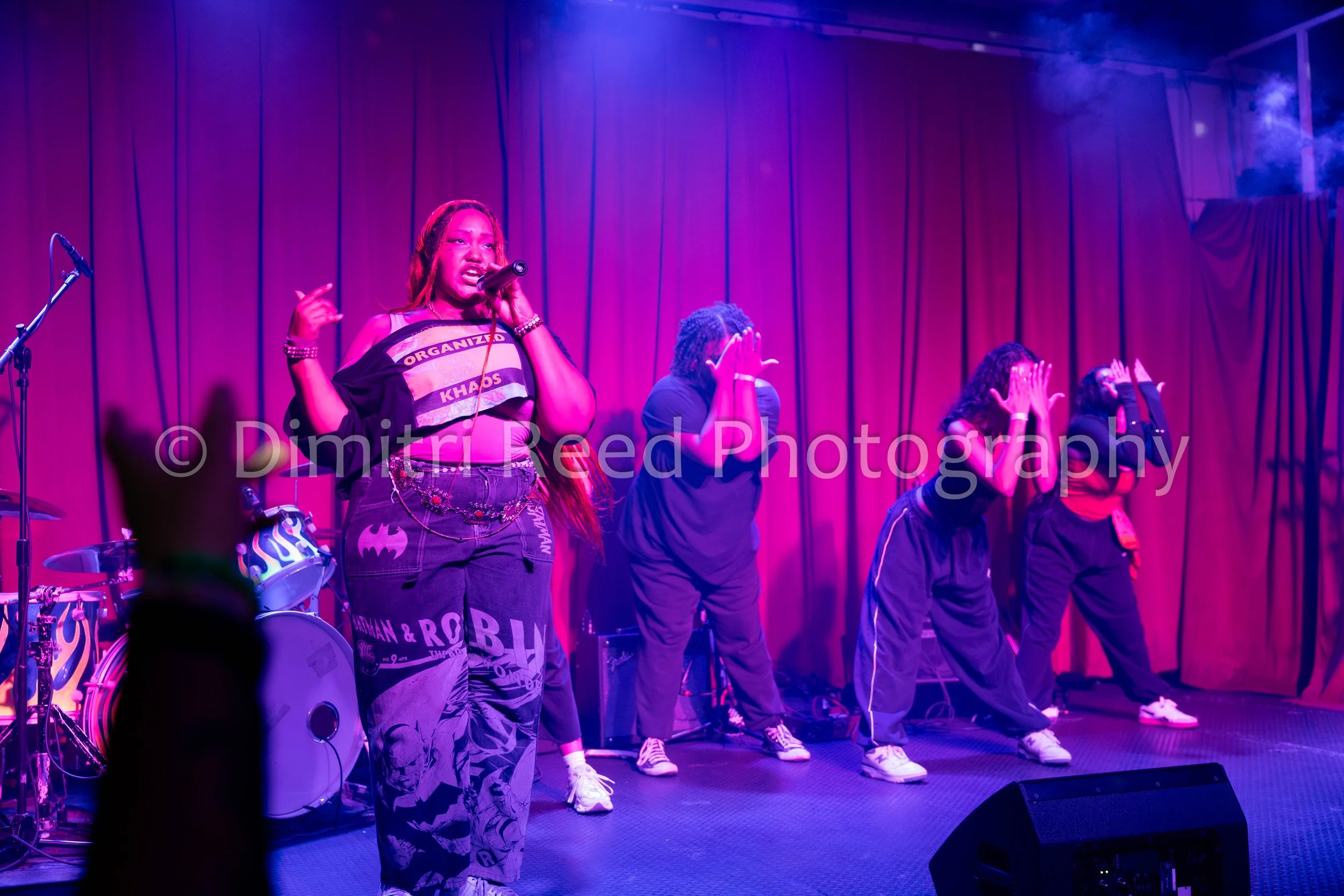 A woman with long red hair singing into a microphone on stage with a group of backup singers behind her, lit with purple stage lighting.