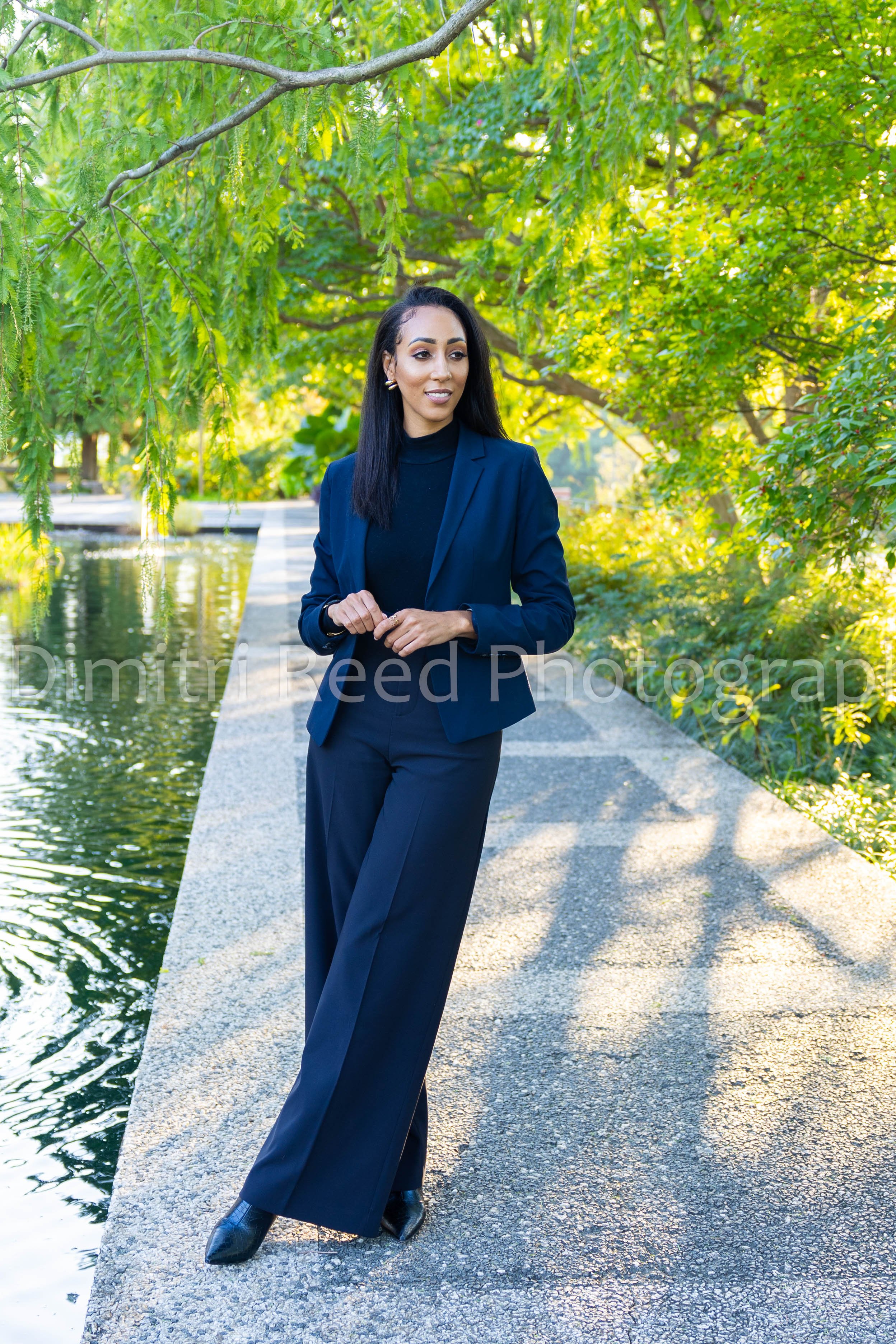 A woman in a navy blue suit and black turtleneck standing on a concrete pathway beside a water feature in a park with green trees overhead.