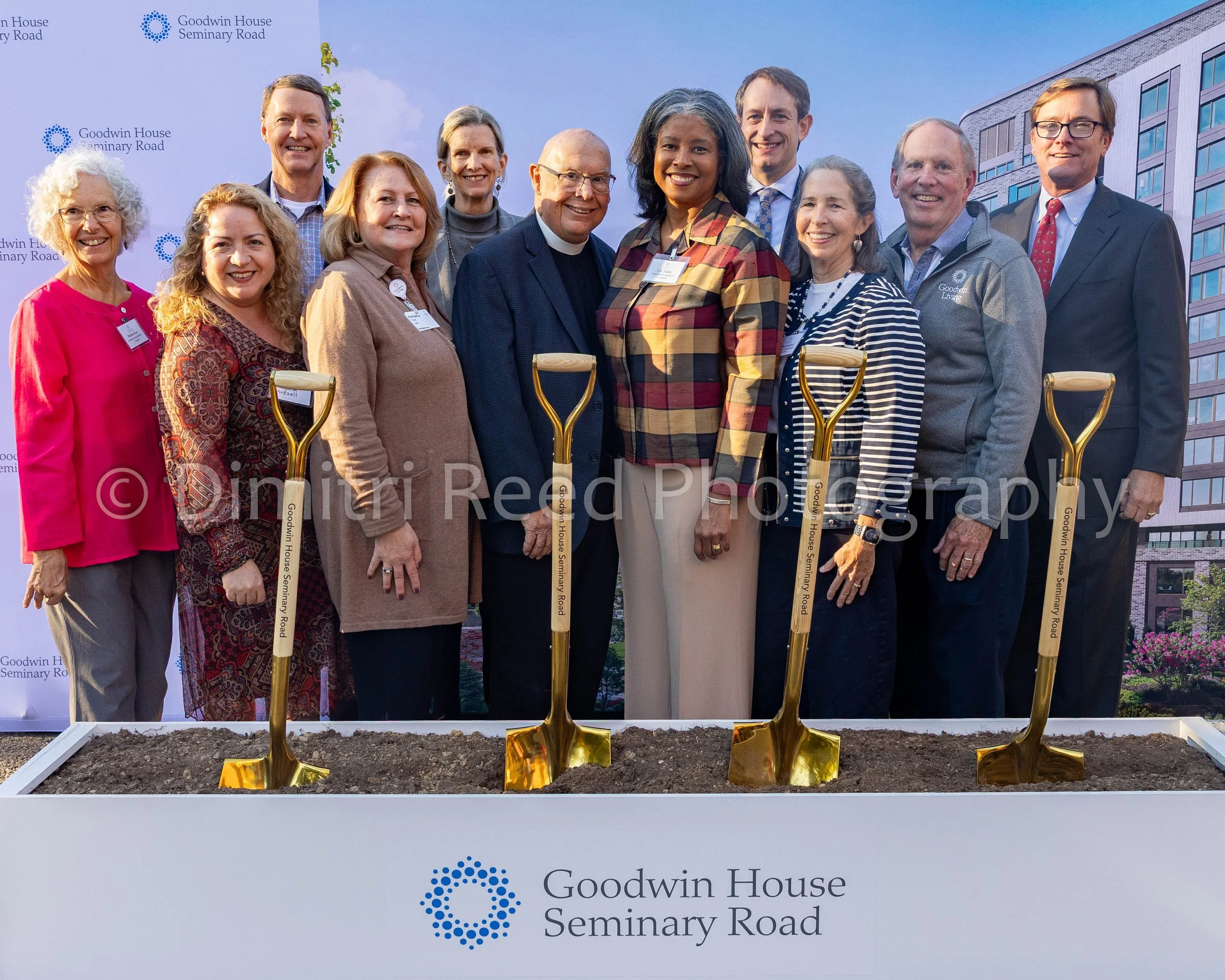 Group of people gathered at a groundbreaking ceremony for Goodwin House Seminary Road, with golden shovels in soil, and a backdrop with the Goodwin House logo and a modern building.