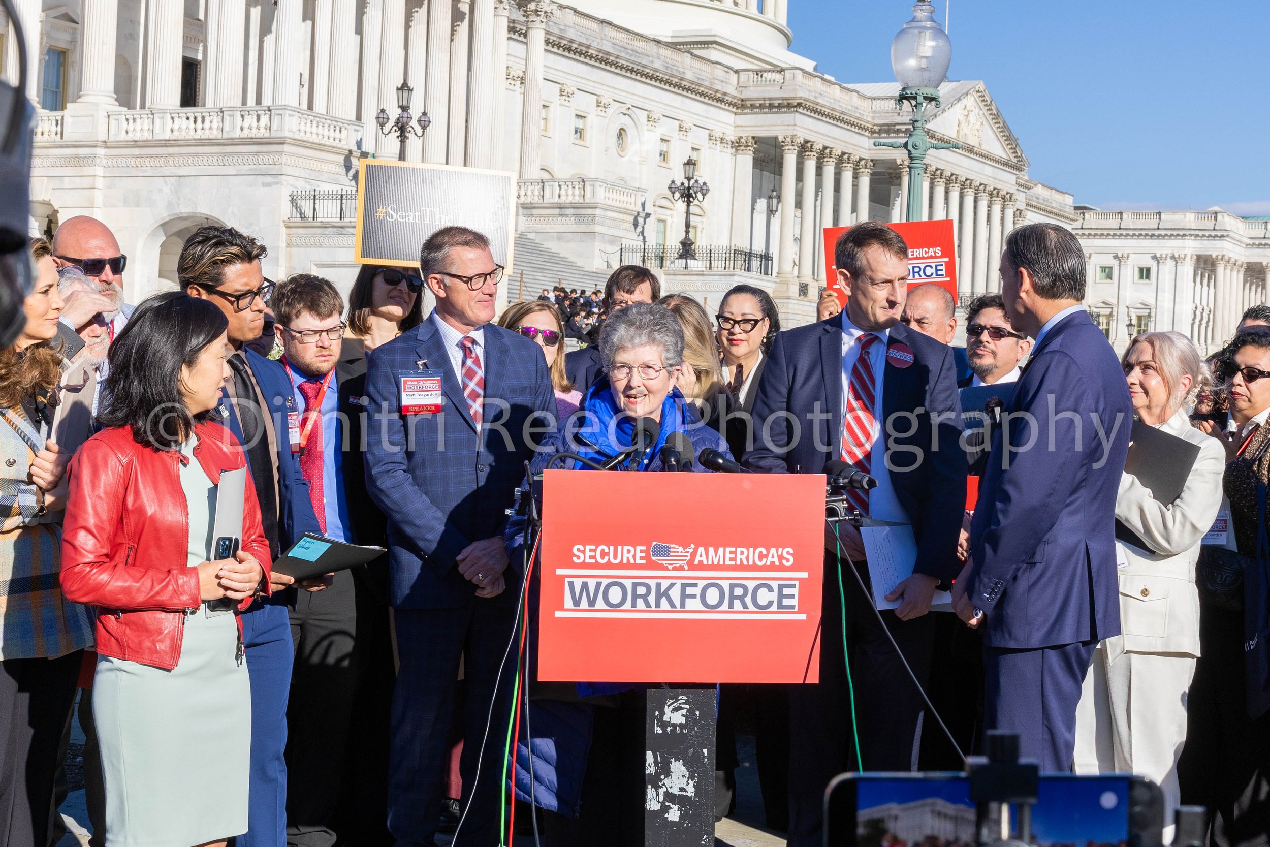 A group of people standing outside near the United States Capitol building, participating in a rally or press conference. There are signs that read 'Secure America's Workforce' and '#SealTheBorder.' The people are dressed in business or casual attire