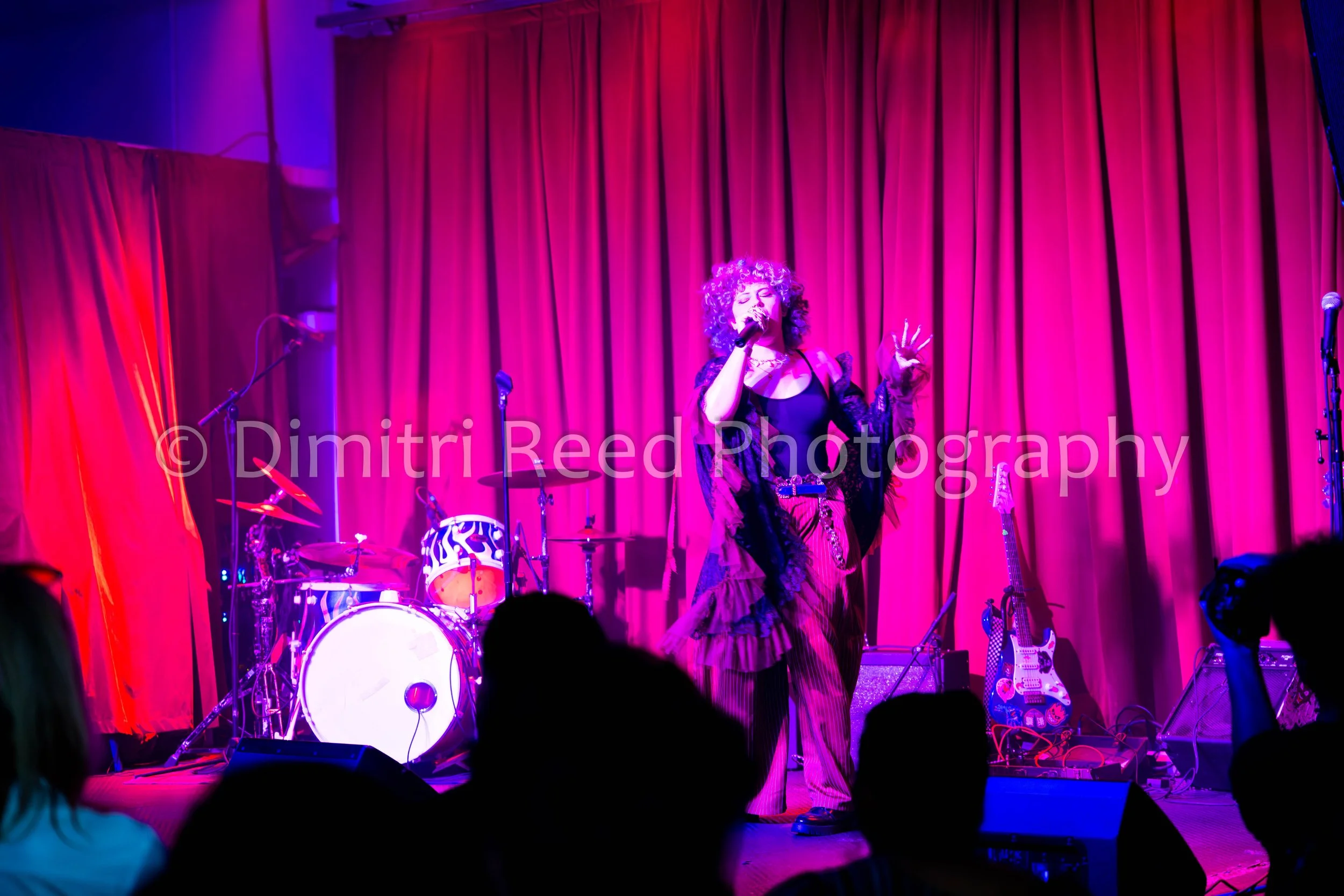 A woman singing on stage with a microphone, surrounded by musical instruments, with red curtains in the background and colored stage lighting.