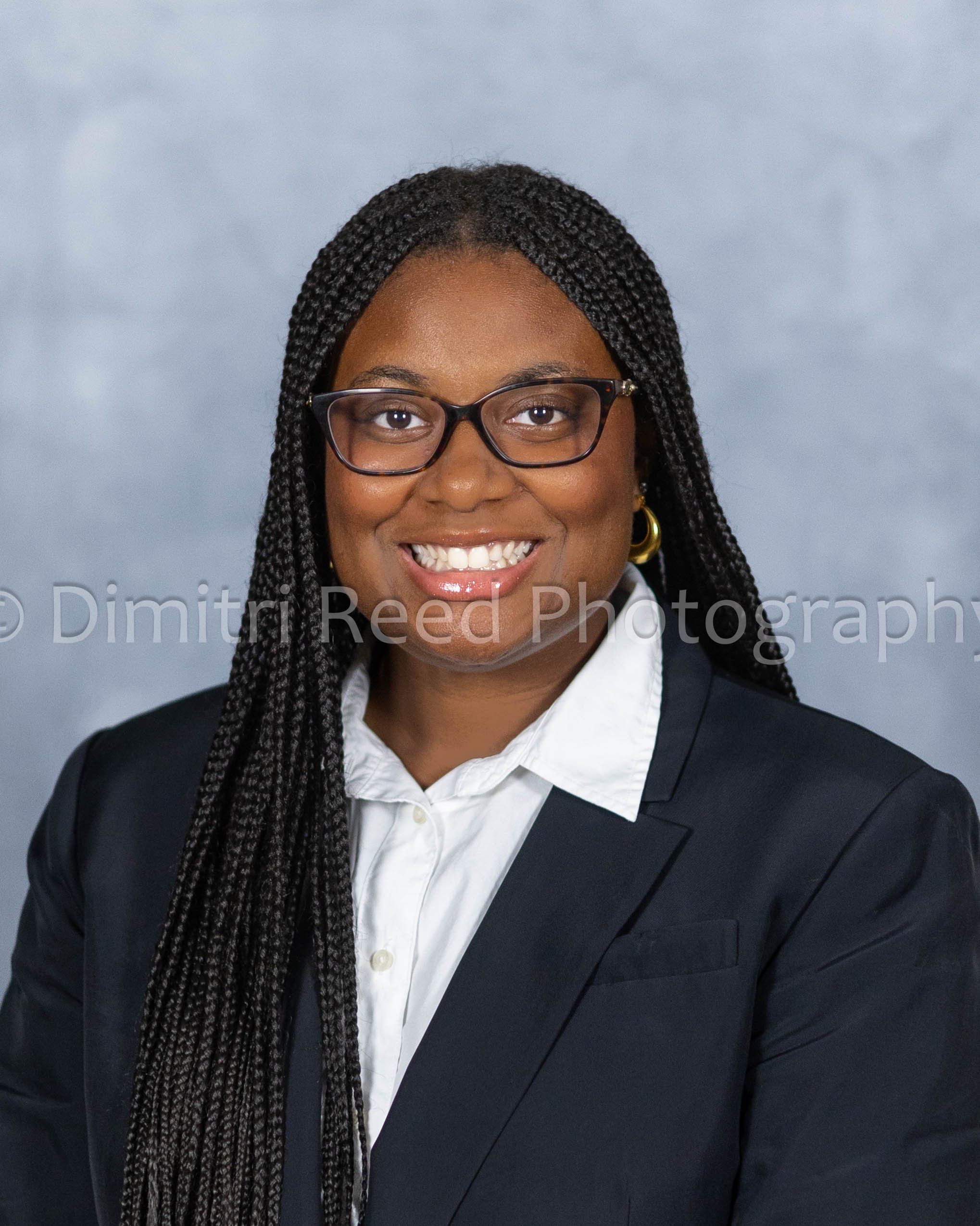 Close-up of an African American woman with braided hair, wearing glasses, a white shirt, and a dark blazer, smiling in front of a gray backdrop.