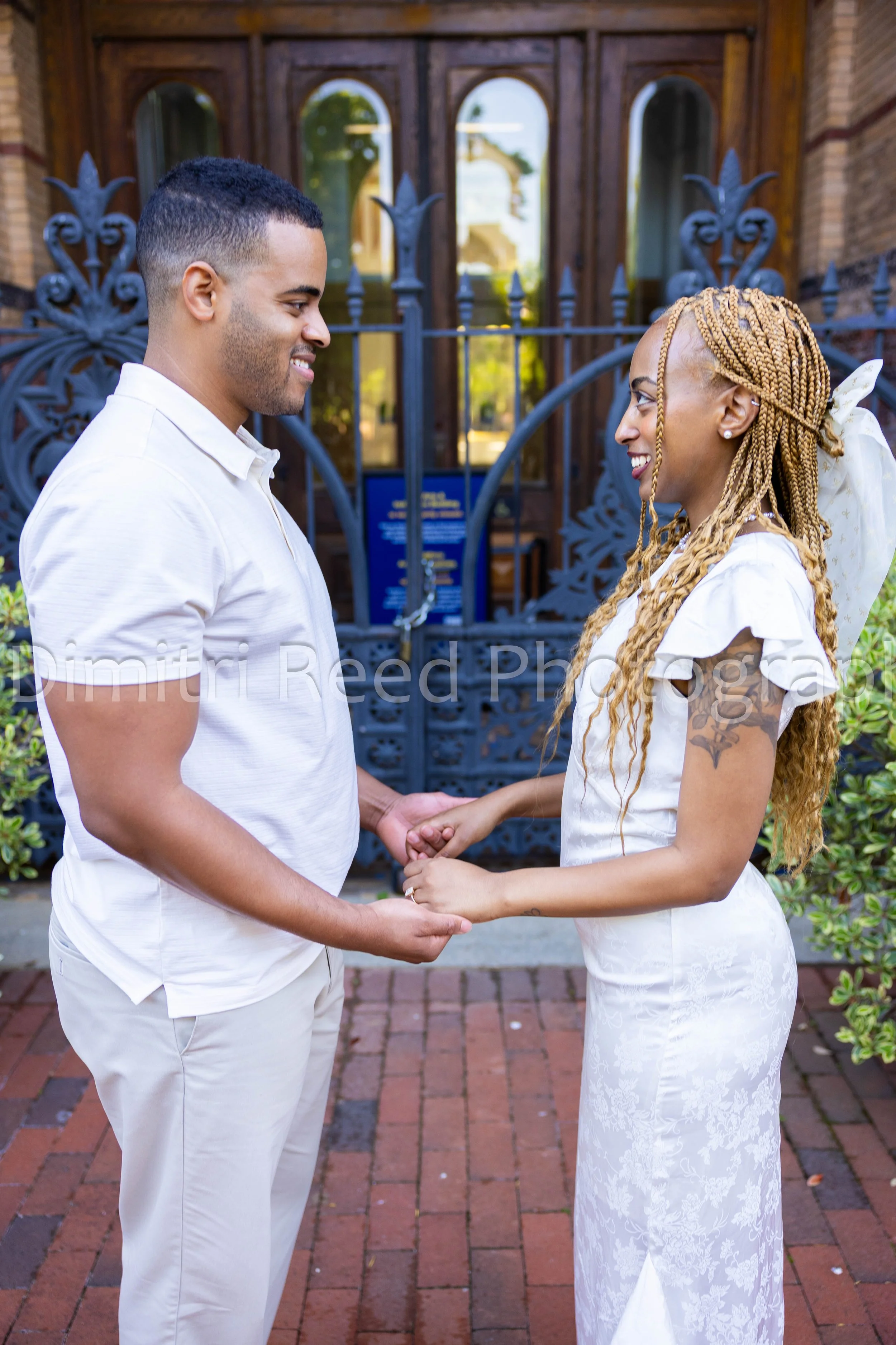 A couple in wedding attire holding hands and looking at each other outside a building with black wrought iron gates and brick walls, smiling.