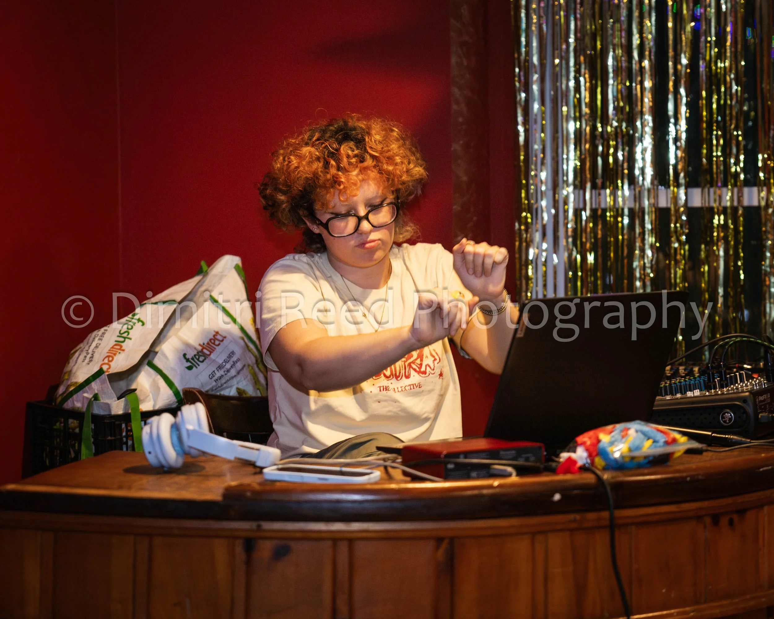 A woman with curly red hair and glasses, wearing a white T-shirt, sits at a table with electronic equipment, cables, and a bag, in front of a gold tinsel backdrop with a red wall behind her.