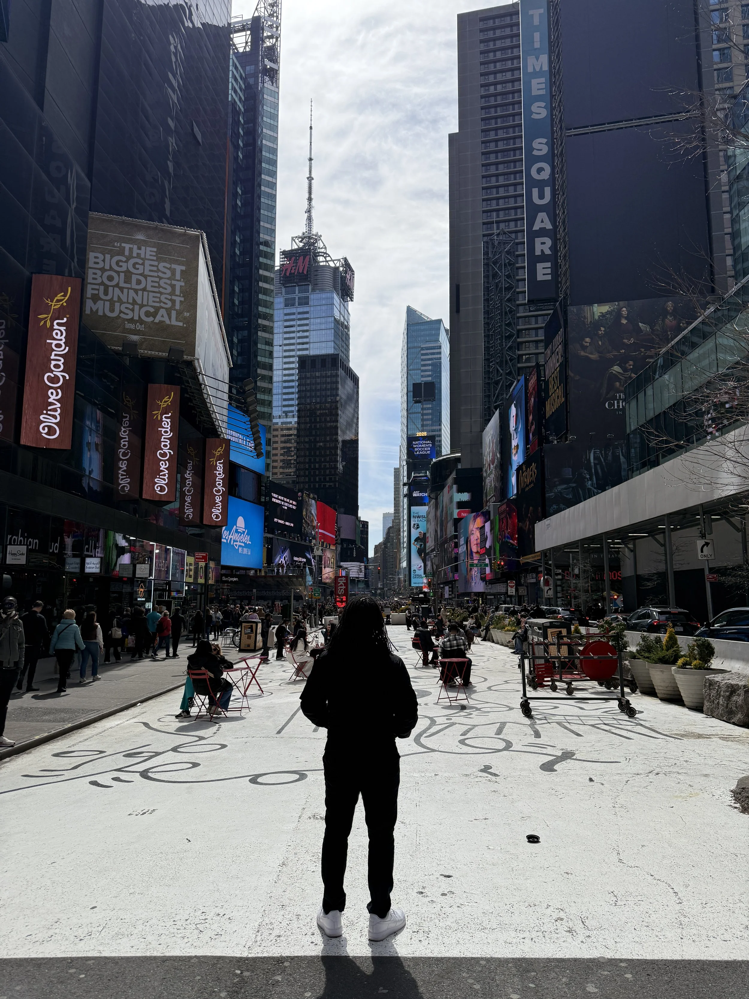 A person with white shoes standing on a painted sidewalk in Times Square, New York City, surrounded by tall buildings, digital billboards, and many people walking and sitting.
