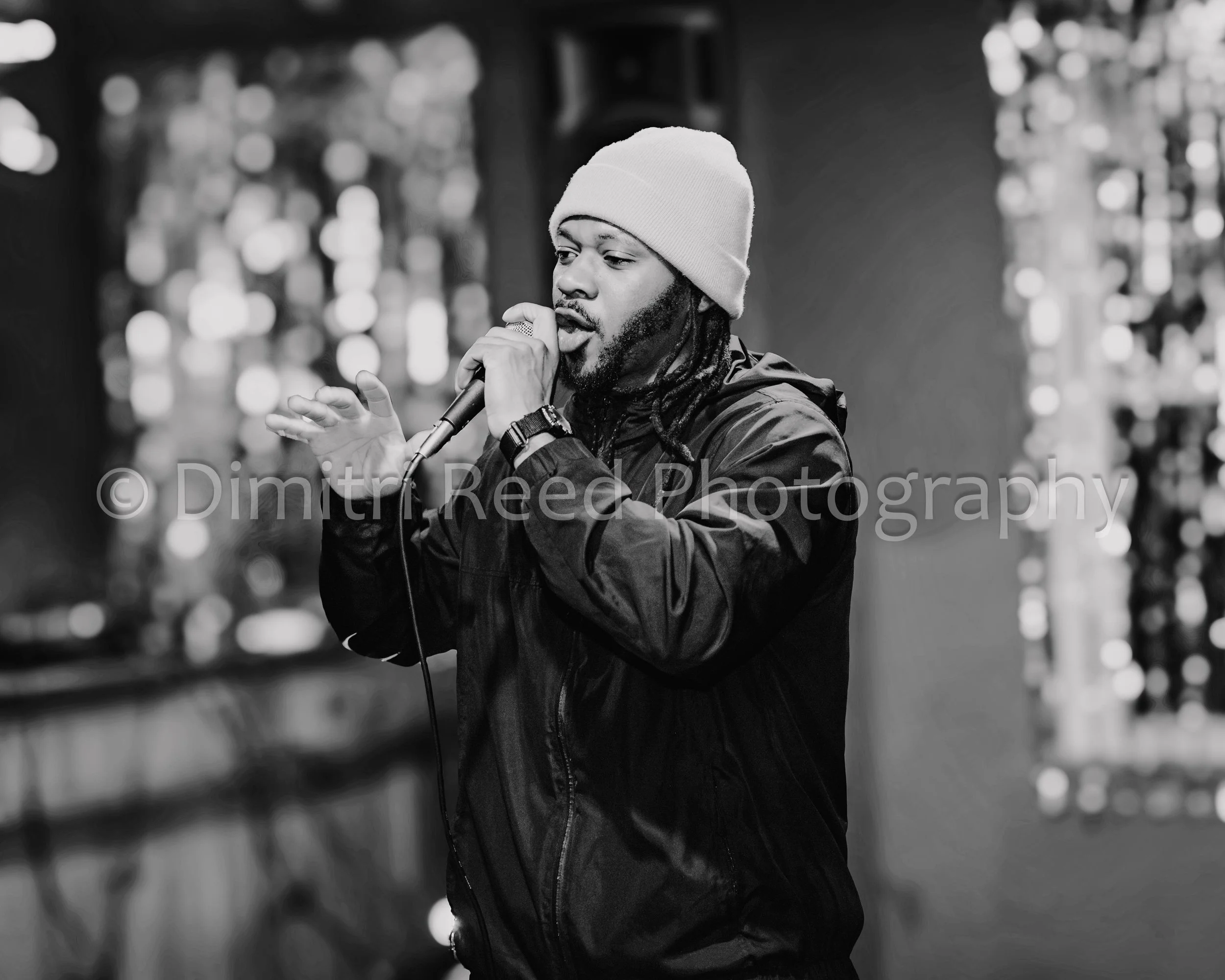 A man with dreadlocks wearing a beanie and a jacket singing into a microphone in a blurred background.