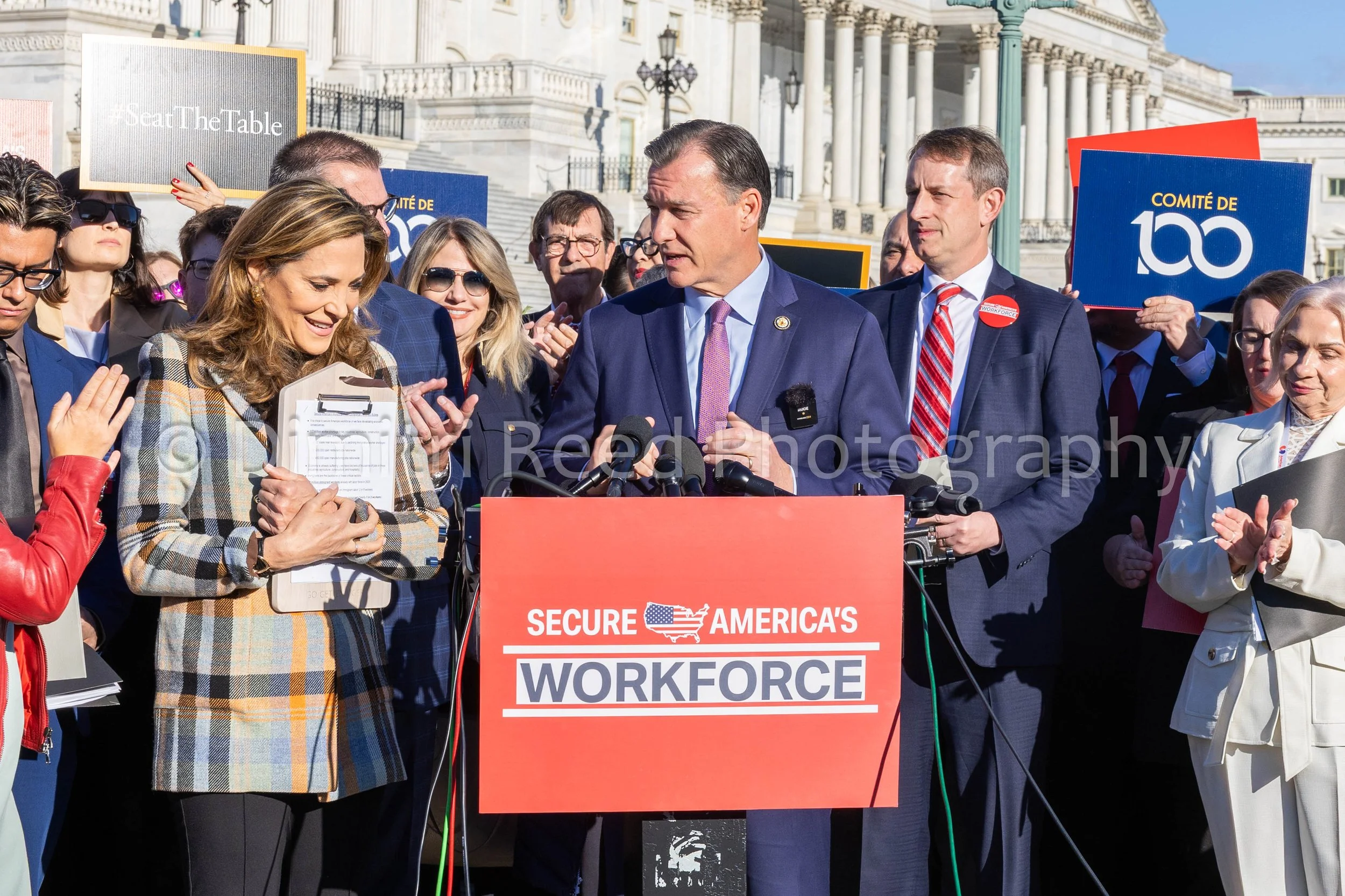 Group of people standing behind a red podium with a sign that reads "Secure America's Workforce" in front of the U.S. Capitol building, some holding signs that say "Join The Table" and "Committee of 100."