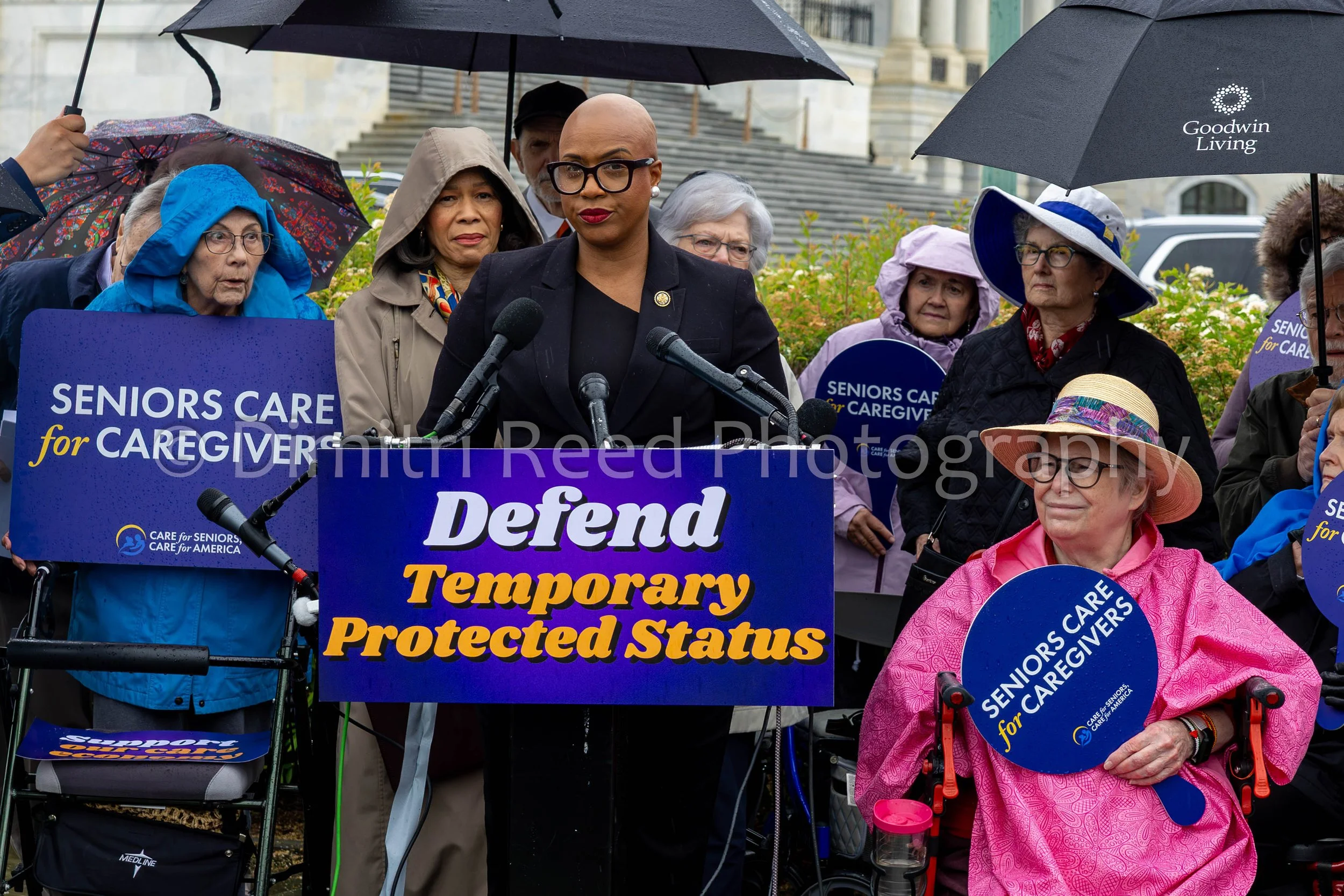A group of senior citizens, some in wheelchairs, holding blue signs and umbrellas, gathered at a protest rally with a speaker at a podium. The signs read 'Seniors Care for Caregivers,' and the large sign in front says 'Defend Temporary Protected Stat