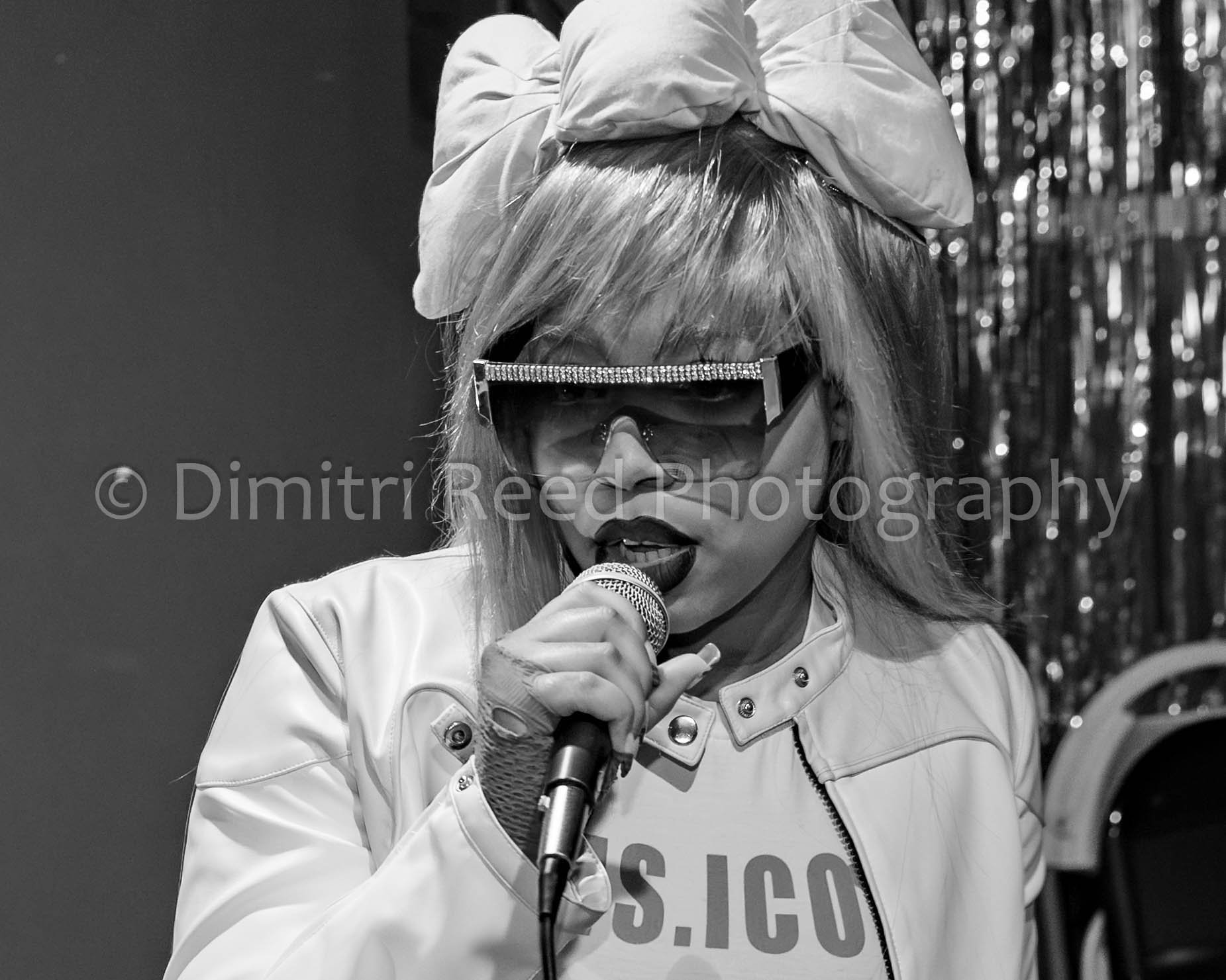 A woman with blonde hair wearing sunglasses, a large bow in her hair, and a white leather jacket, singing into a microphone. She is in front of a shiny, reflective background, possibly on a stage.