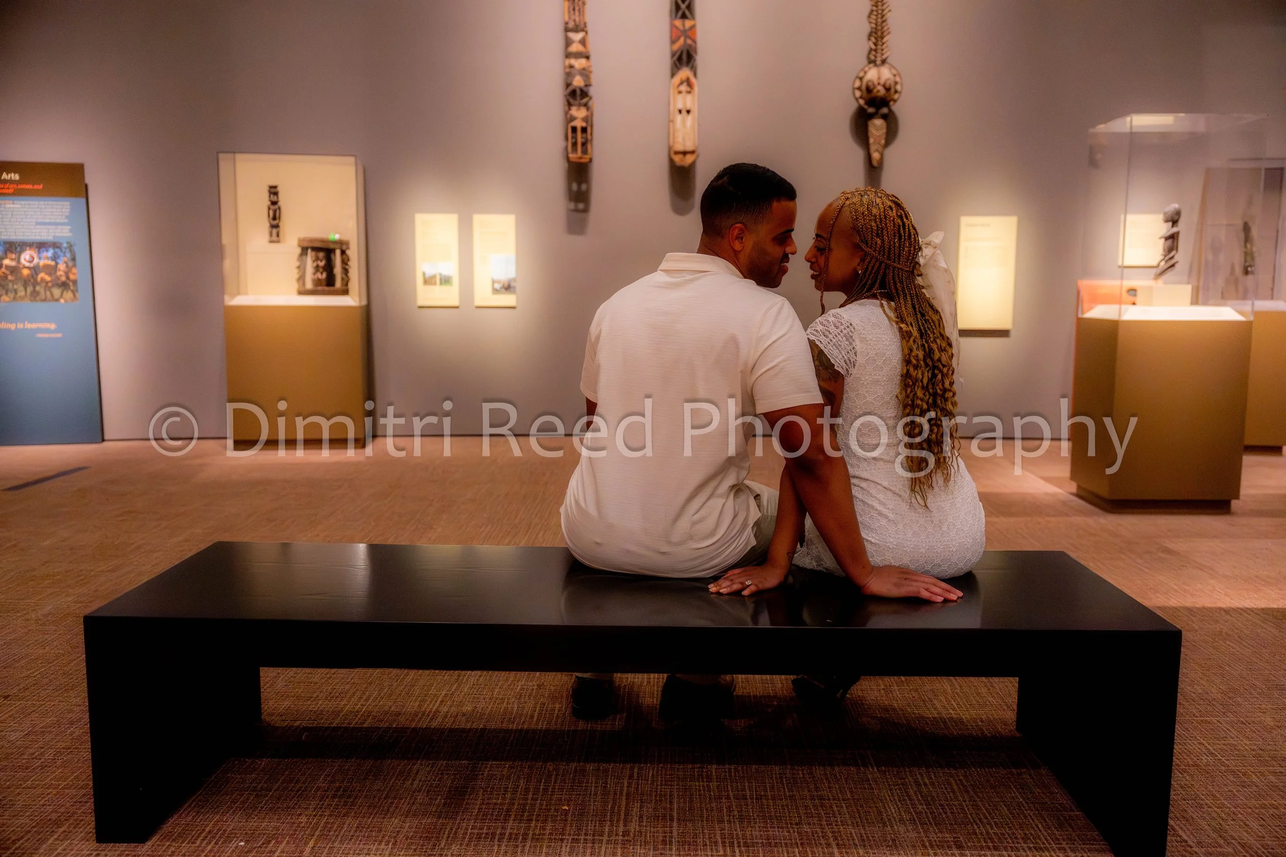 A couple is sitting on a black bench in an art museum, touching foreheads and looking at each other, with Native American cultural artifacts hanging on the wall behind them.