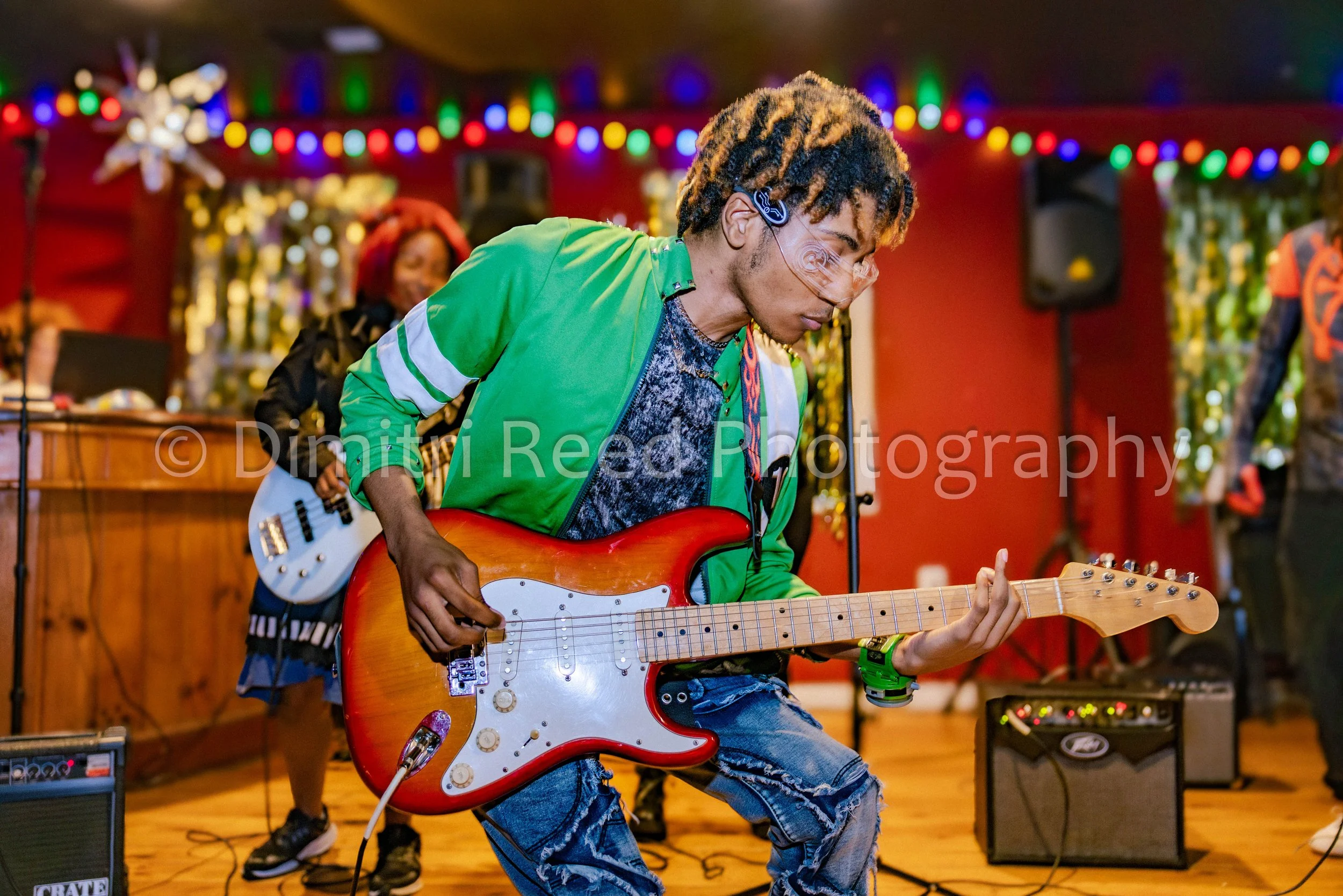 A young man with dreadlocks wearing glasses and a green jacket playing an electric guitar at a decorated indoor event with colorful lights and music equipment.