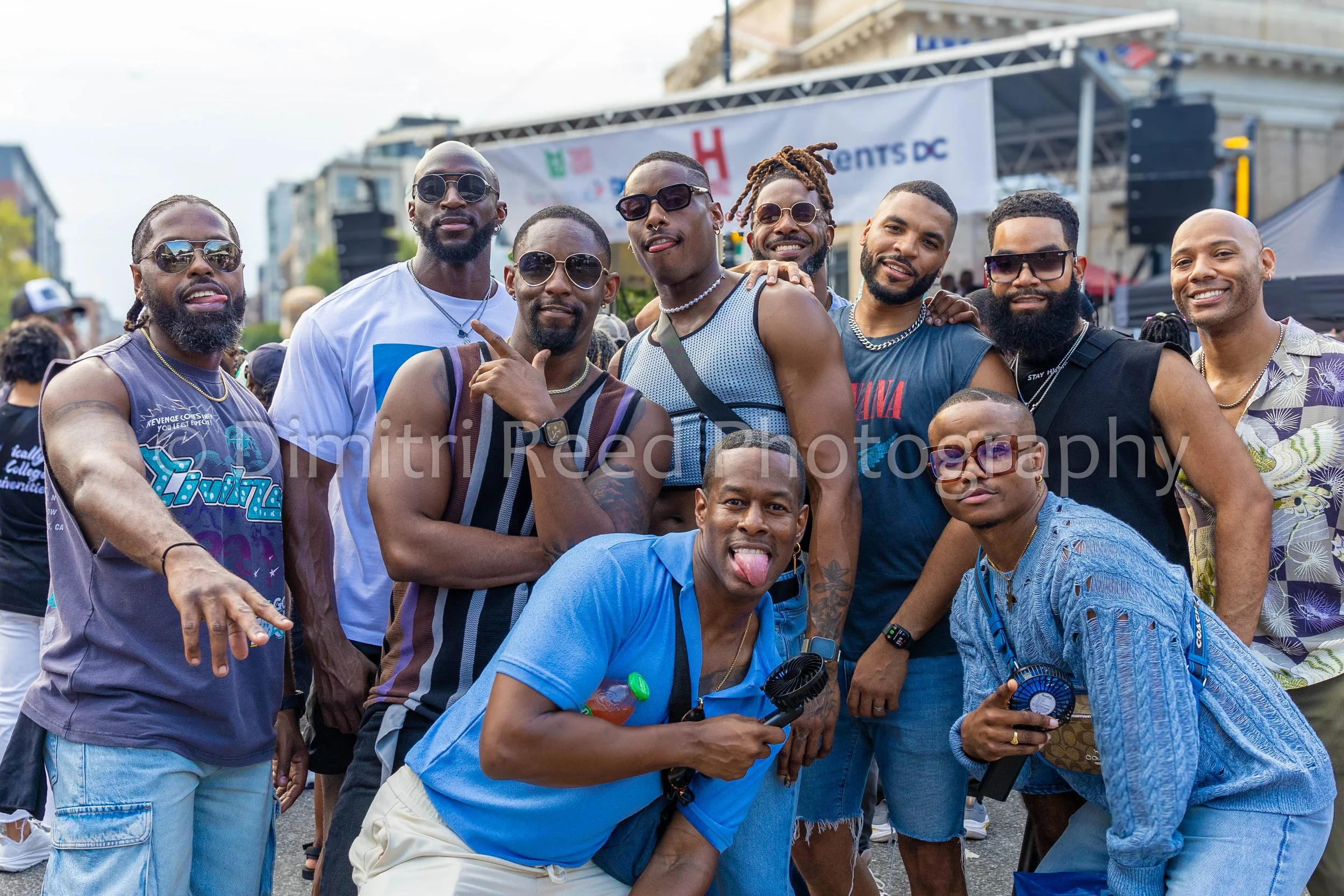 Group of men in casual clothing and sunglasses posing in front of an outdoor stage at a public event.