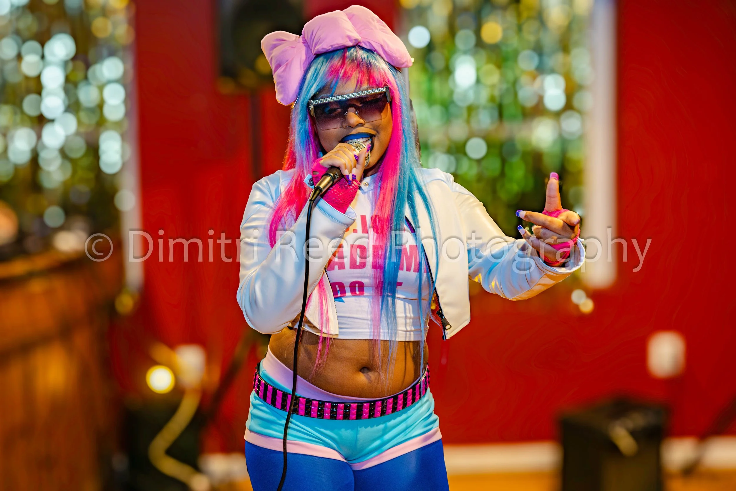 A woman with long multicolored hair, wearing large sunglasses with rhinestones, a pink bow headband, a white cropped varsity jacket, a pink and white T-shirt, pink gloves, blue shorts with a pink and black checkered waistband, and blue tights, singin