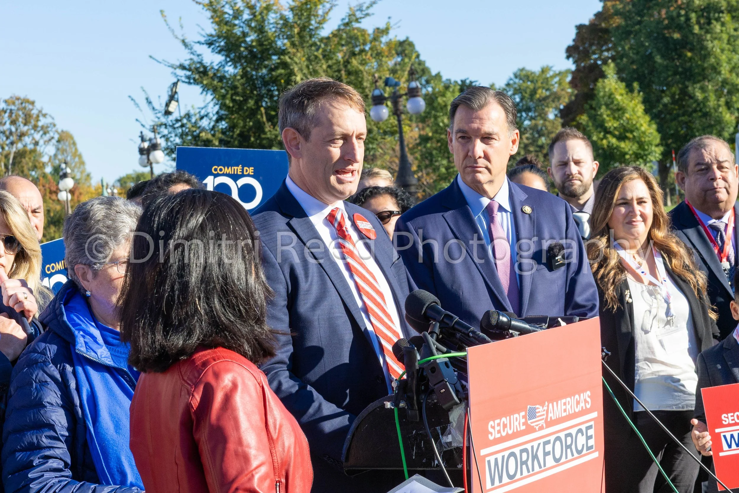 A group of people gathered outdoors for a public event, with two men in suits speaking at a podium with microphones. The background shows trees and a clear sky, and there are signs that say 'Secure America's Workforce' and 'Committee of 100'.