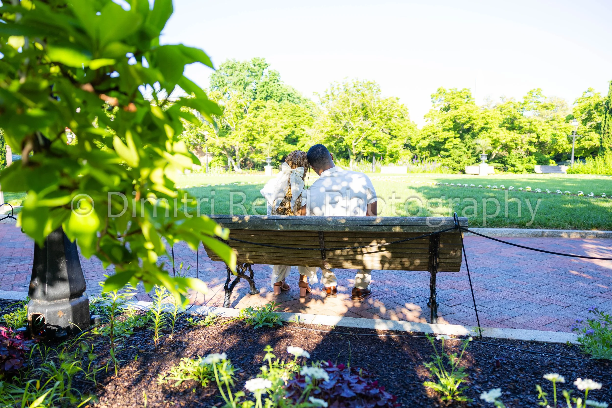 A couple sitting on a bench in a park, surrounded by green trees and flowers, with bright sunlight.
