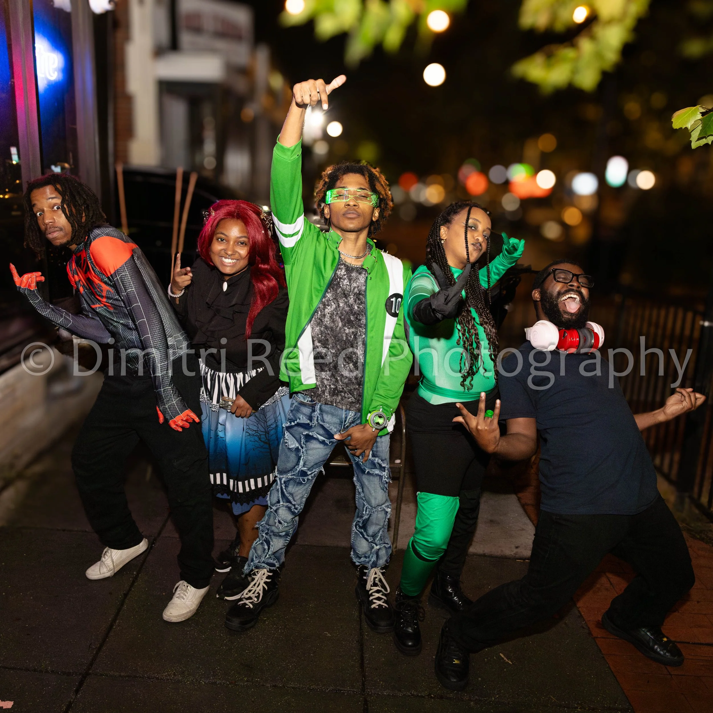 A group of six people, dressed in colorful and casual clothing, standing on a wet sidewalk at night. They are posing happily, some making gestures with their hands, with blurred city lights in the background.