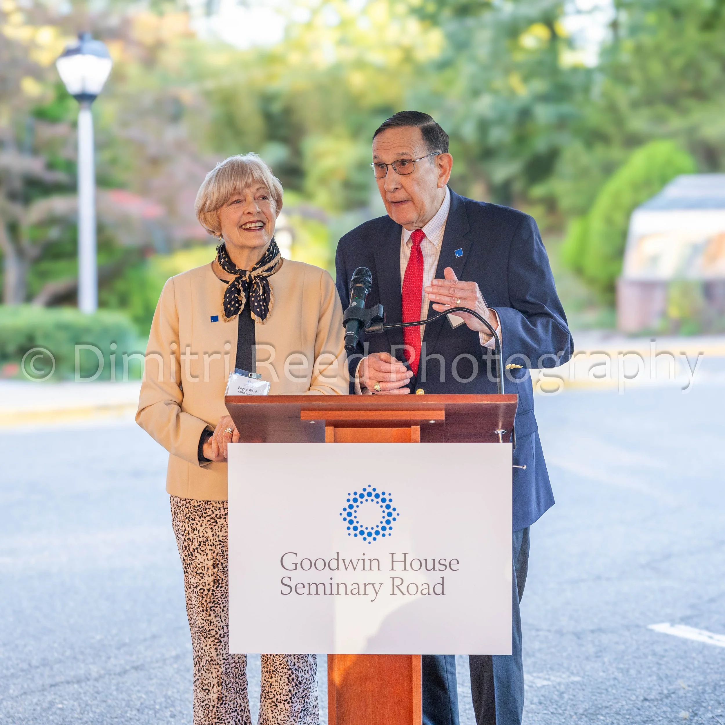 Two people, an elderly woman and an elderly man, standing behind a podium at an outdoor event. The woman is smiling and wearing a beige jacket with a polka dot scarf, and the man is speaking into a microphone, dressed in a navy suit with a red tie. T