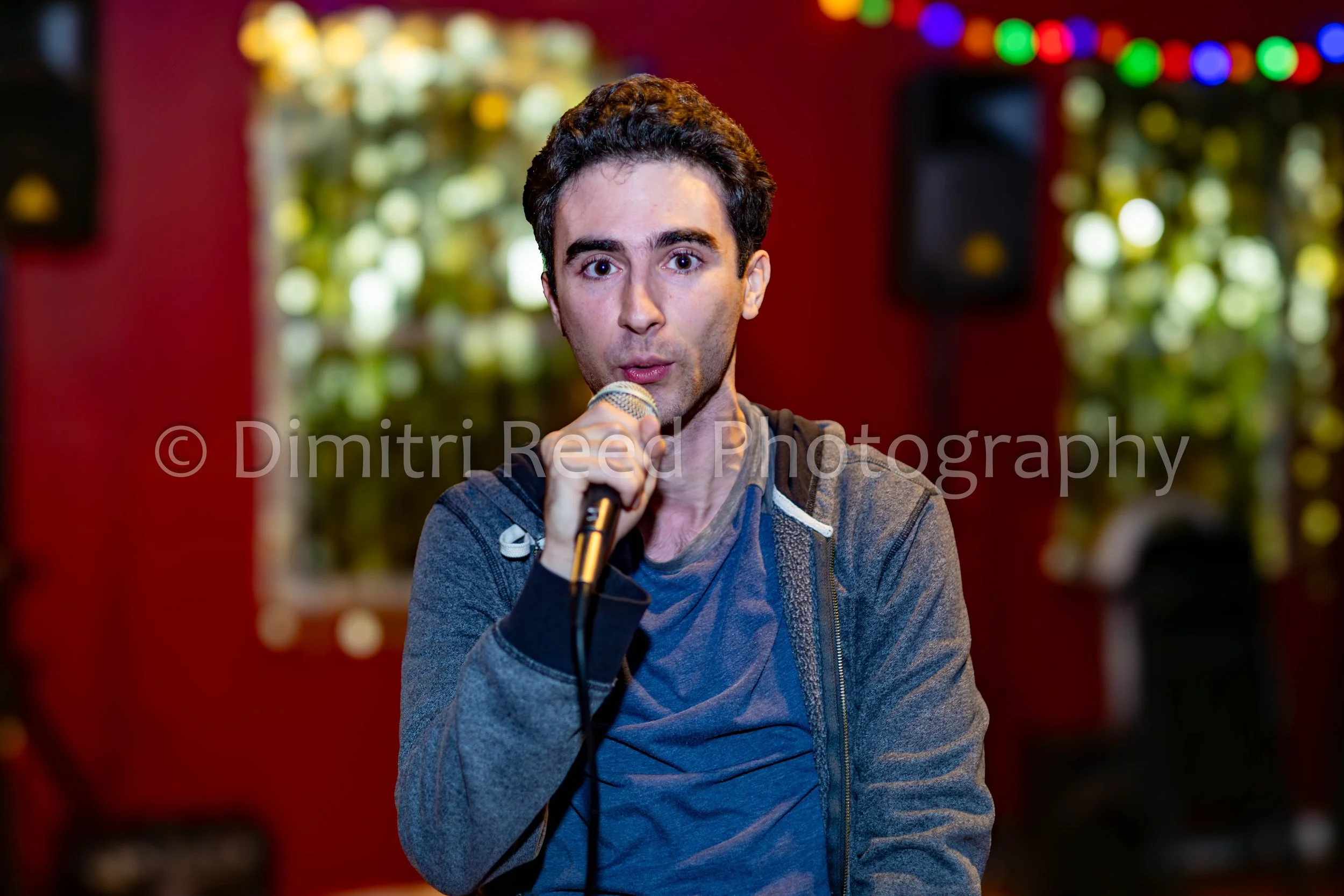 A man with dark hair, wearing a gray jacket, holding a microphone, speaking indoors against a background of colorful, blurred Christmas lights.