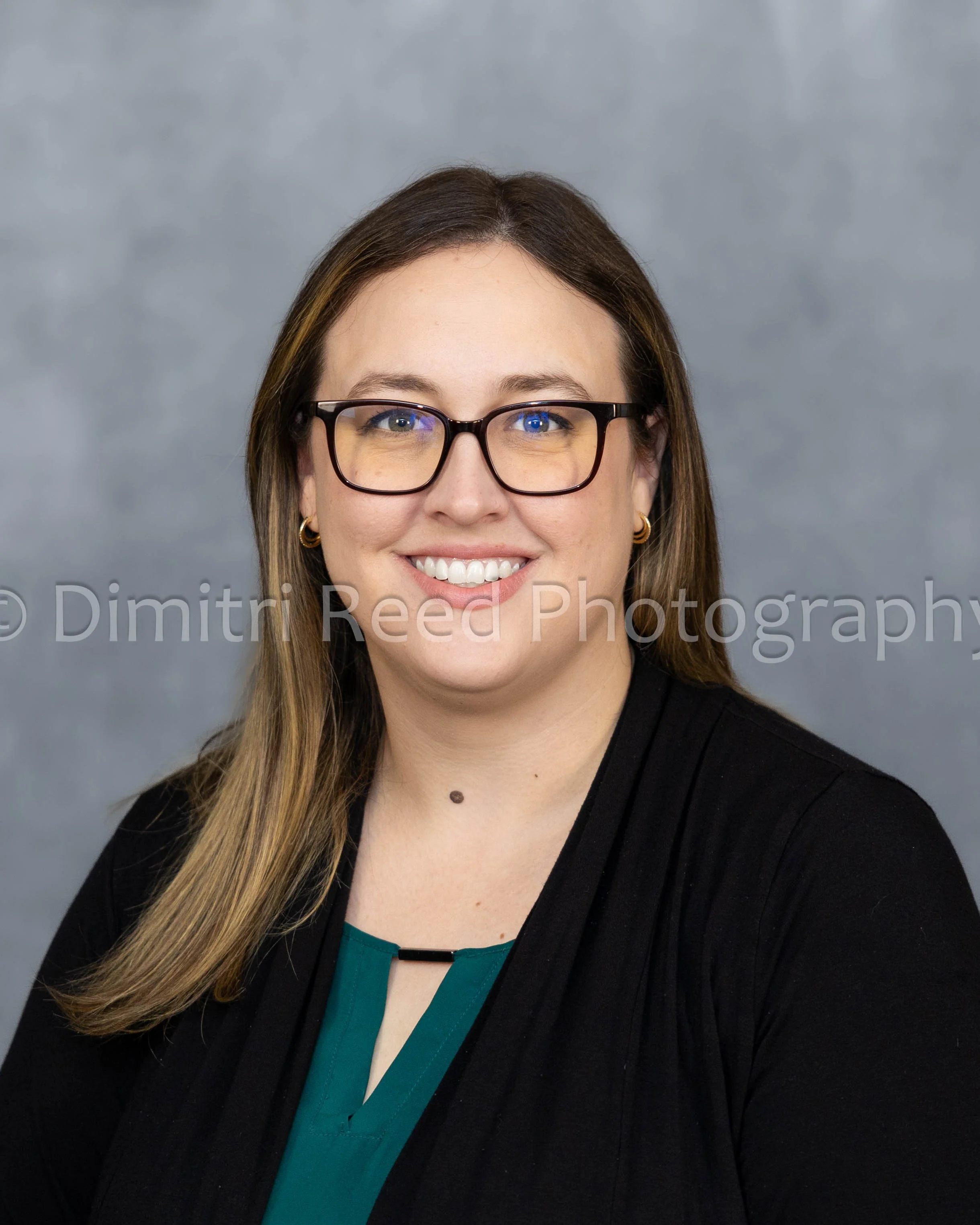 A woman with long brown hair, smiling, wearing glasses, a black blazer, and a teal blouse, sitting in front of a gray background.