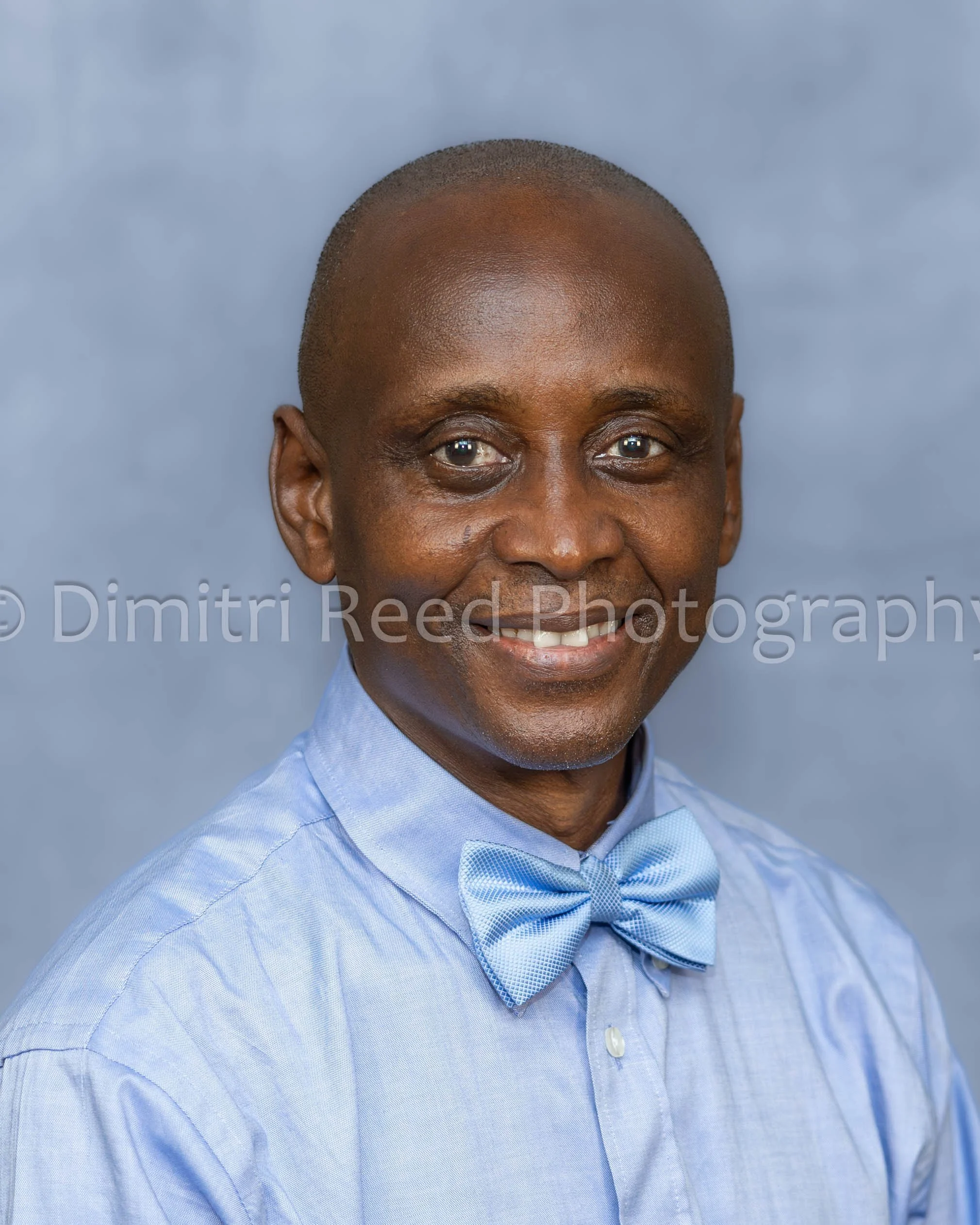 Close-up of a smiling African American man wearing a light blue dress shirt and matching bow tie, with a neutral gray background.