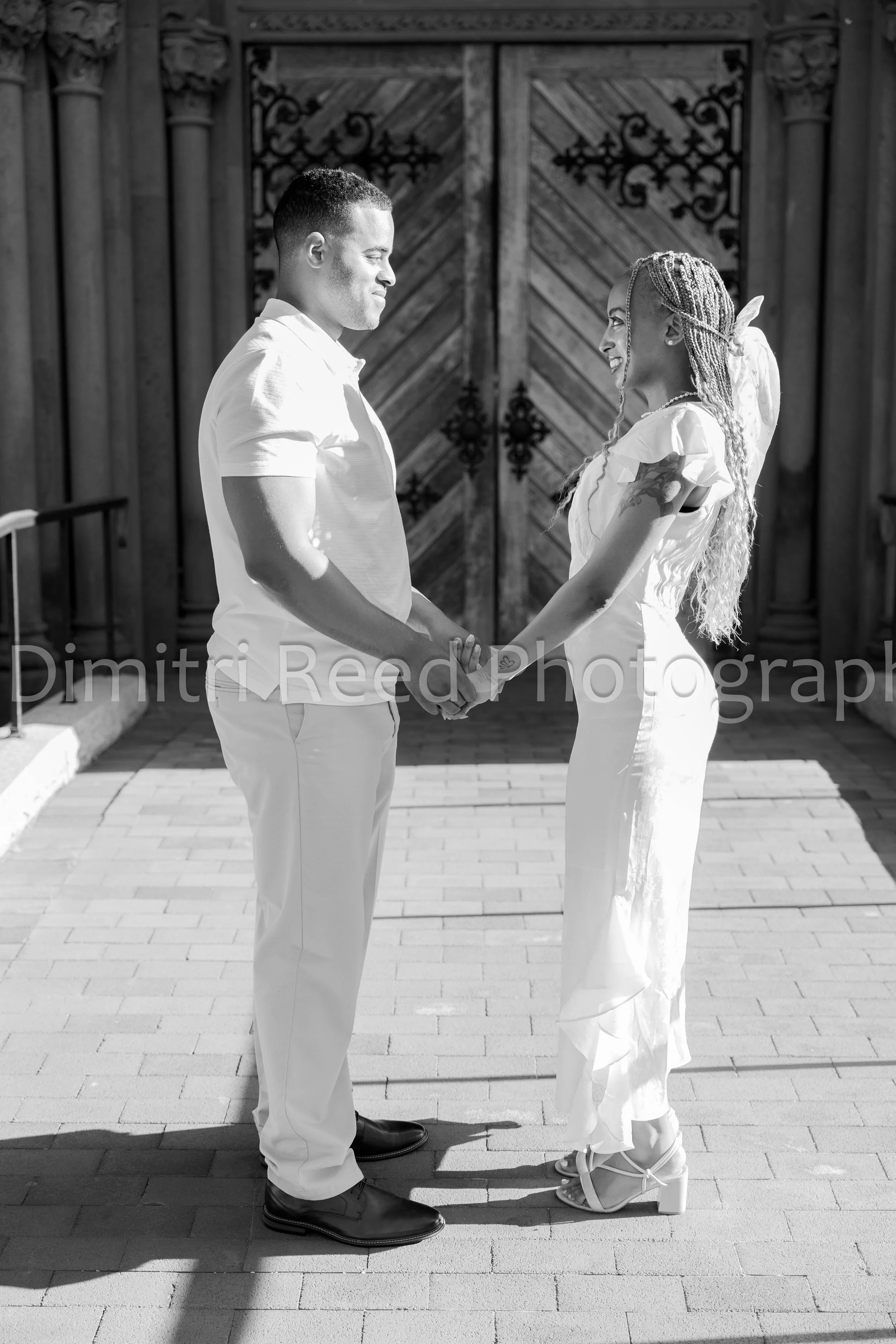 A man and woman holding hands and smiling at each other, standing outdoors in front of a large wooden door with wrought iron details, both dressed in white, with the woman wearing white heels and the man wearing black shoes.