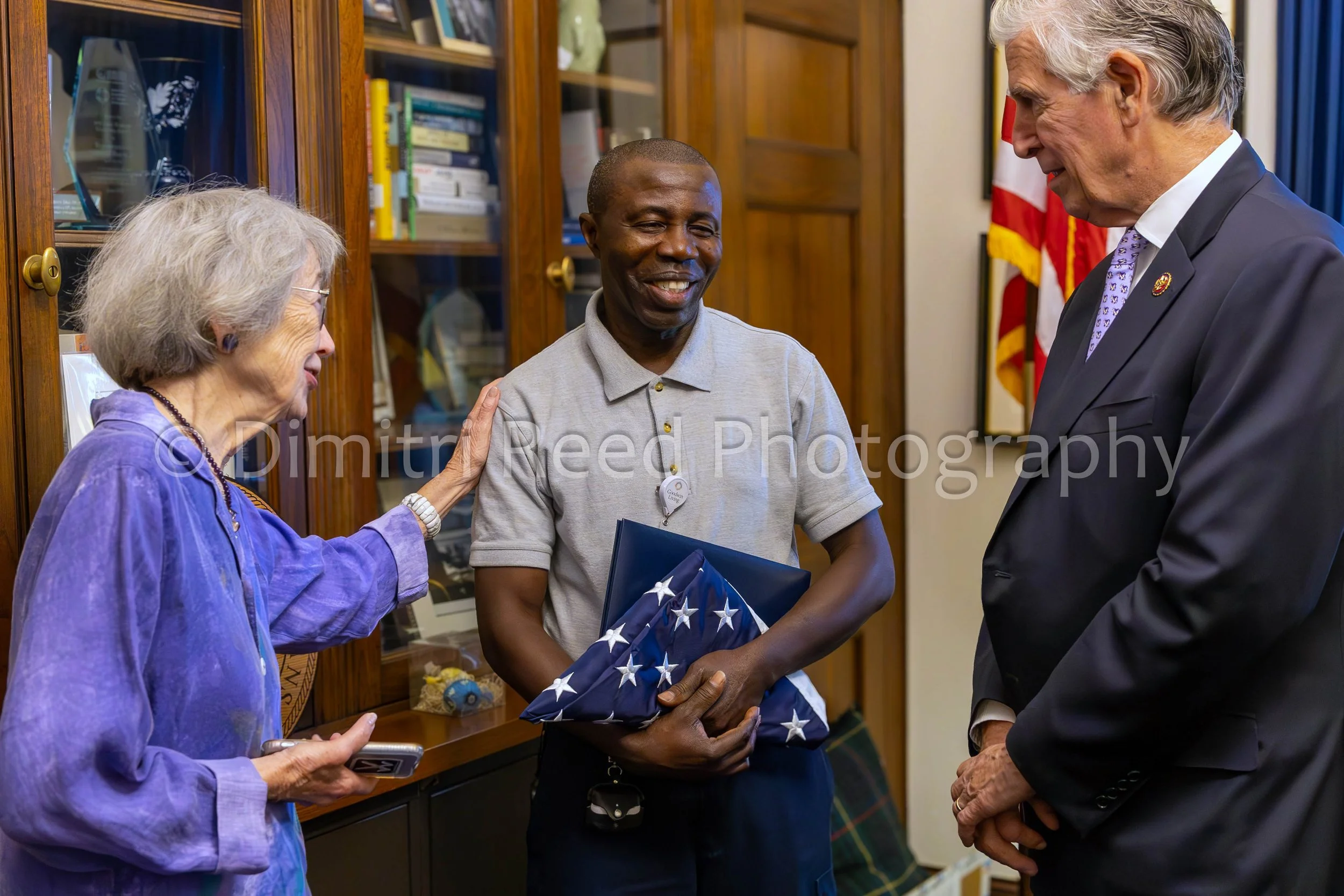 Four people engaged in conversation in a room with wooden bookshelves. One woman is wearing a purple top, another man is holding a folded American flag, and another man is wearing a dark suit. An older woman is touching the man holding the flag. An A