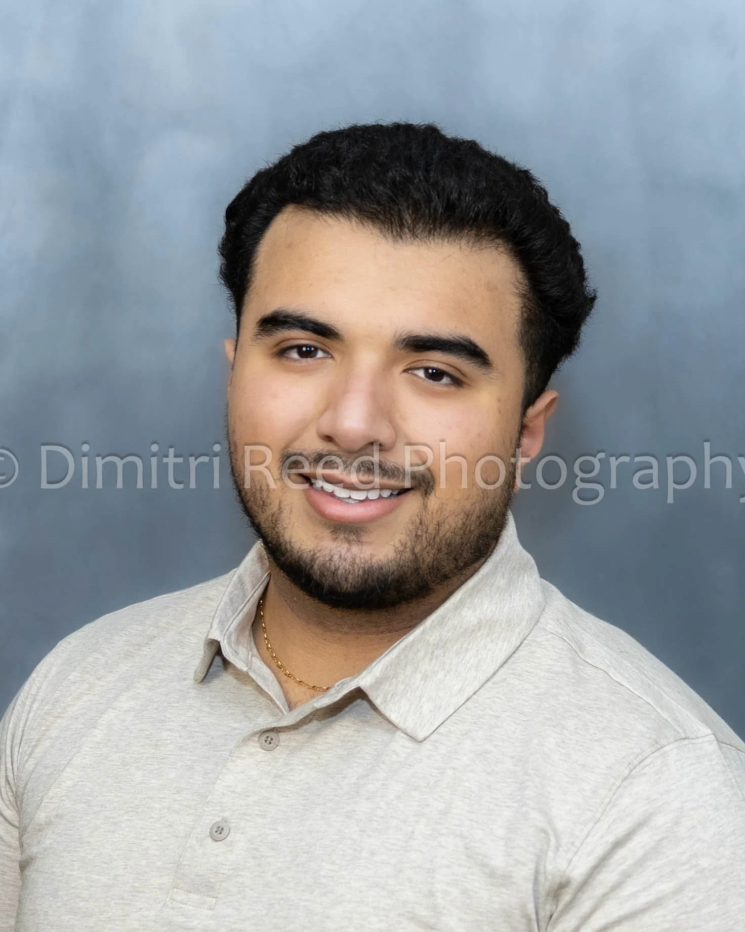 A young man with dark hair and a beard is smiling, wearing a light beige collared shirt and a gold chain, standing in front of a blue background.