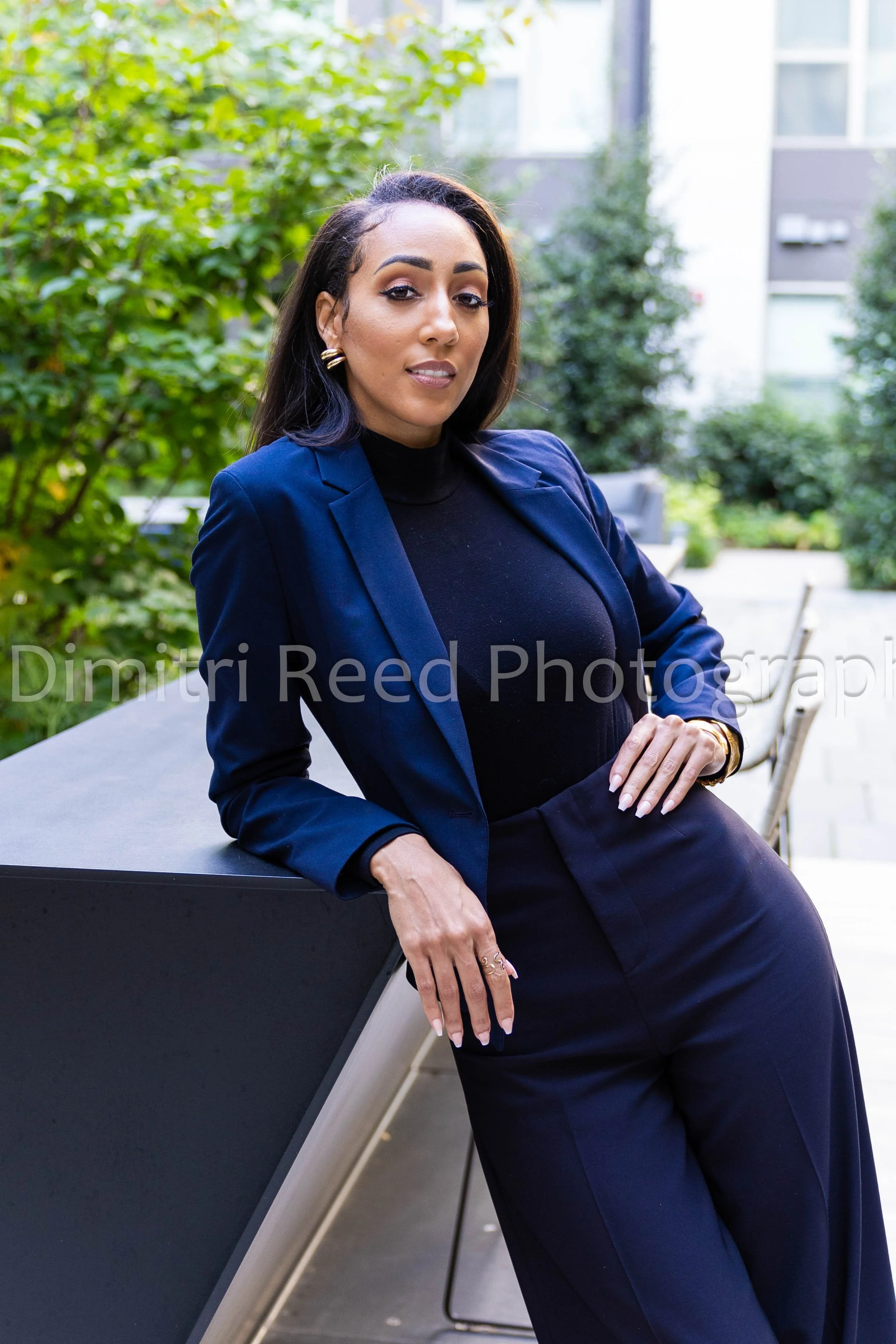 A confident woman with dark hair, wearing a navy blazer and black top, posing outdoors with greenery and modern buildings in the background.