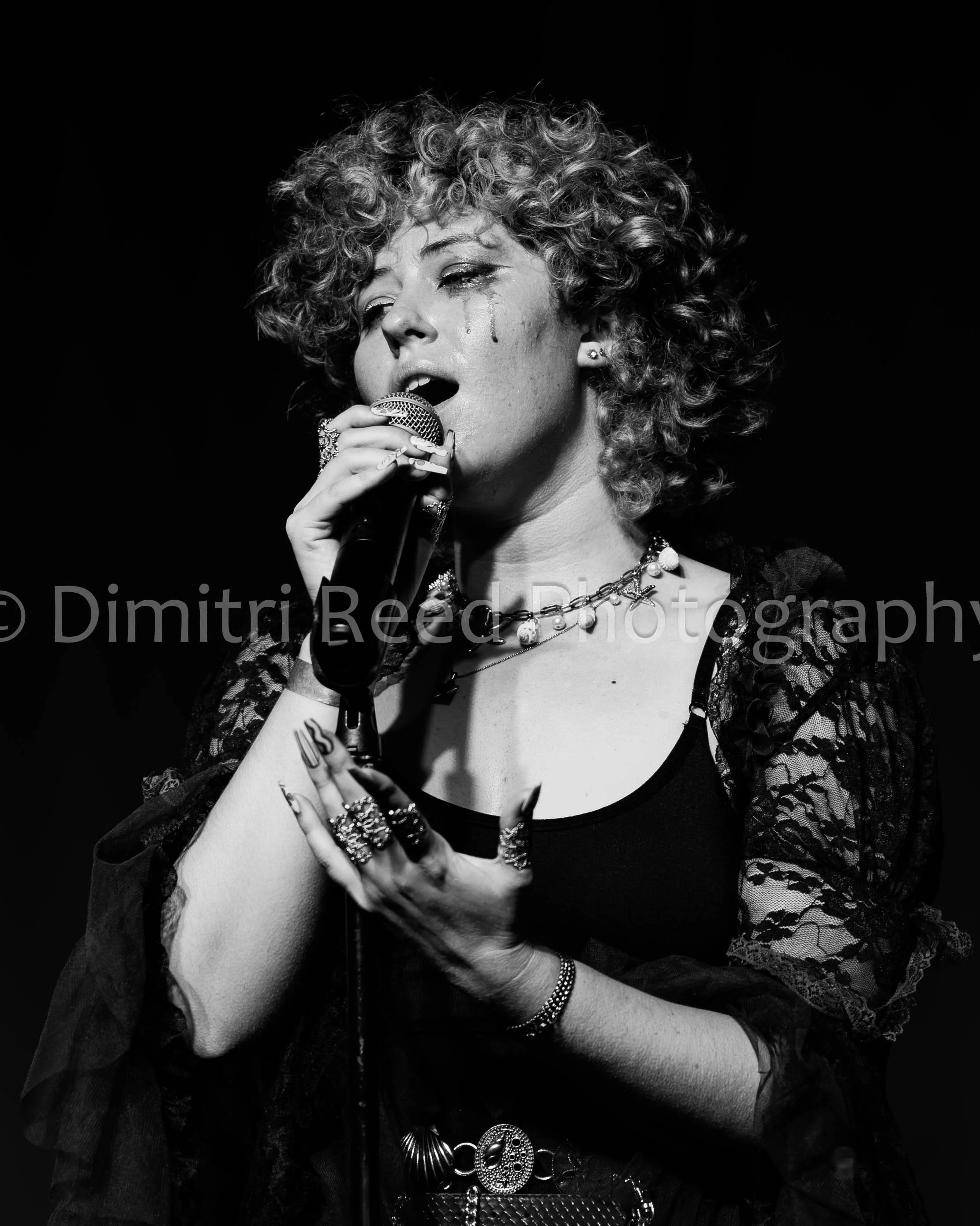 Black and white photo of a woman singing into a microphone with eyes partially closed, wearing lace clothing, multiple jewelry including rings, earrings, necklaces, and bracelets, with curly hair and teary eyes.