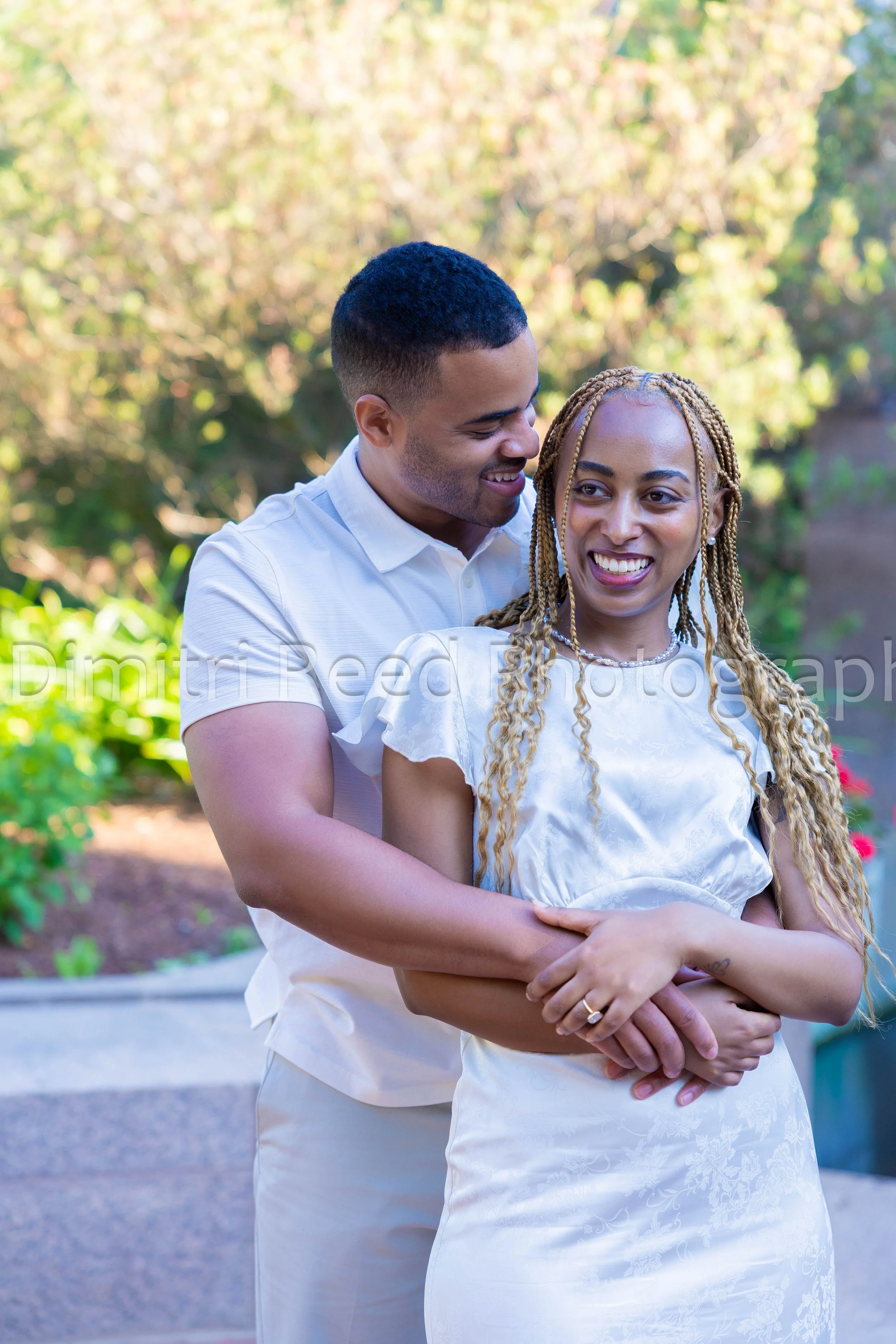 A couple embracing outdoors on a sunny day, smiling and looking at each other. The man is wearing a white shirt, and the woman is wearing a white dress. The background features green foliage and sunlight.