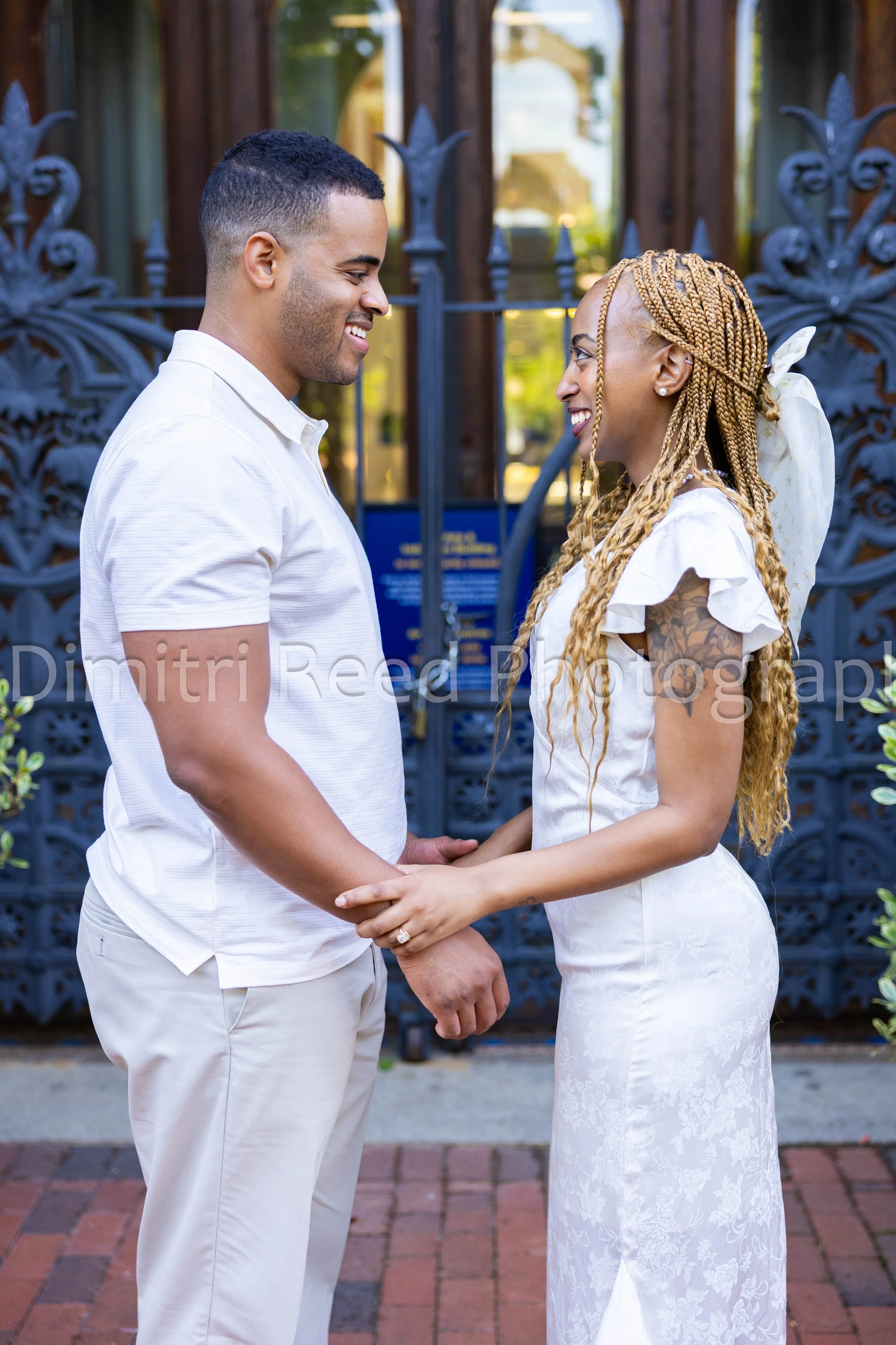 A couple holding hands, standing face to face outdoors on a brick sidewalk, with a decorative gate and greenery in the background. The man is wearing a white shirt and pants, smiling with closed eyes; the woman in a white dress with floral patterns, 