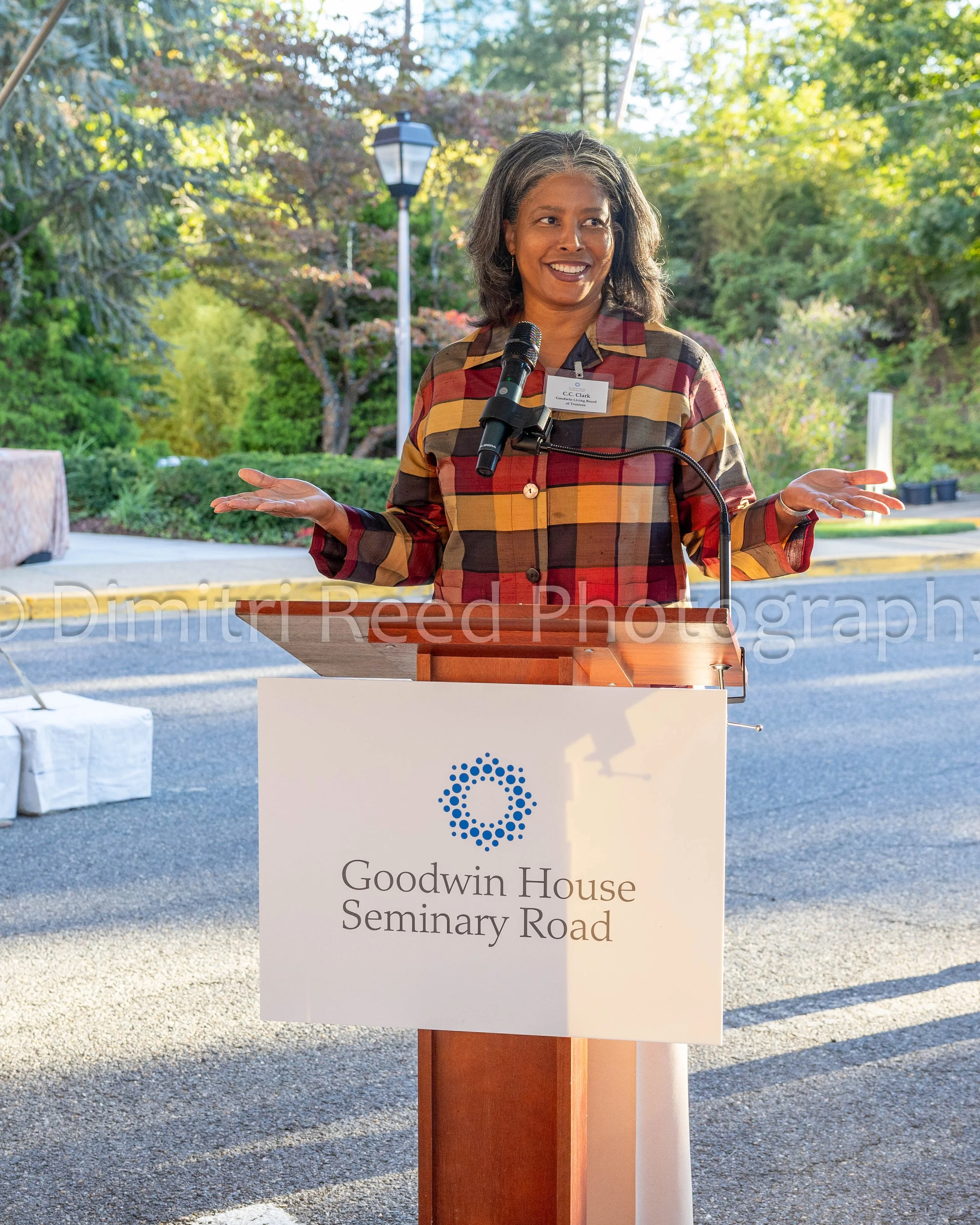 A woman with gray hair wearing a plaid shirt standing outdoors with trees, shrubs, and a streetlamp in the background, speaking at a podium with a sign that reads 'Goodwin House Seminary Road'.