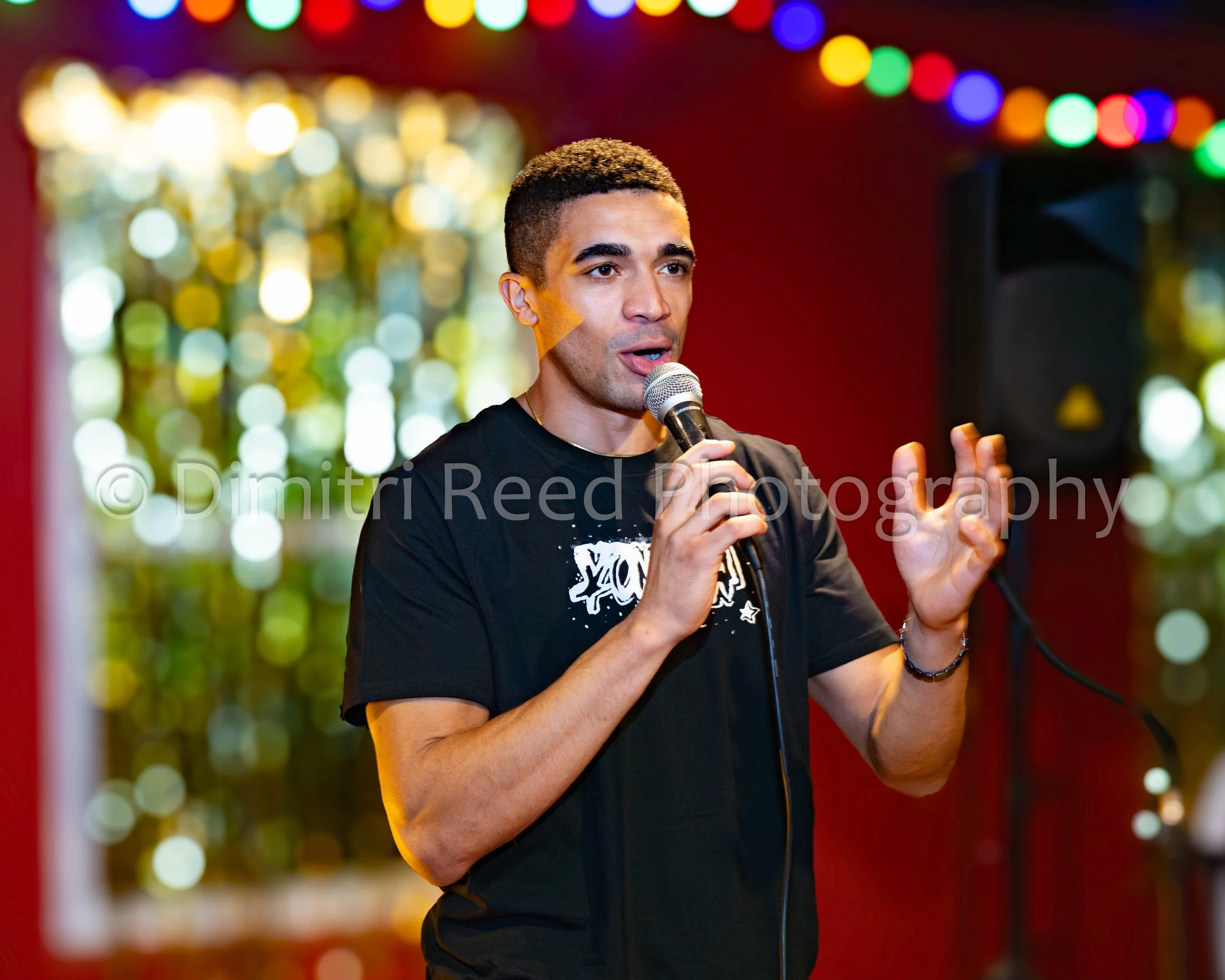 A young man with short curly hair singing into a microphone at a party or performance, with colorful blurred lights in the background.