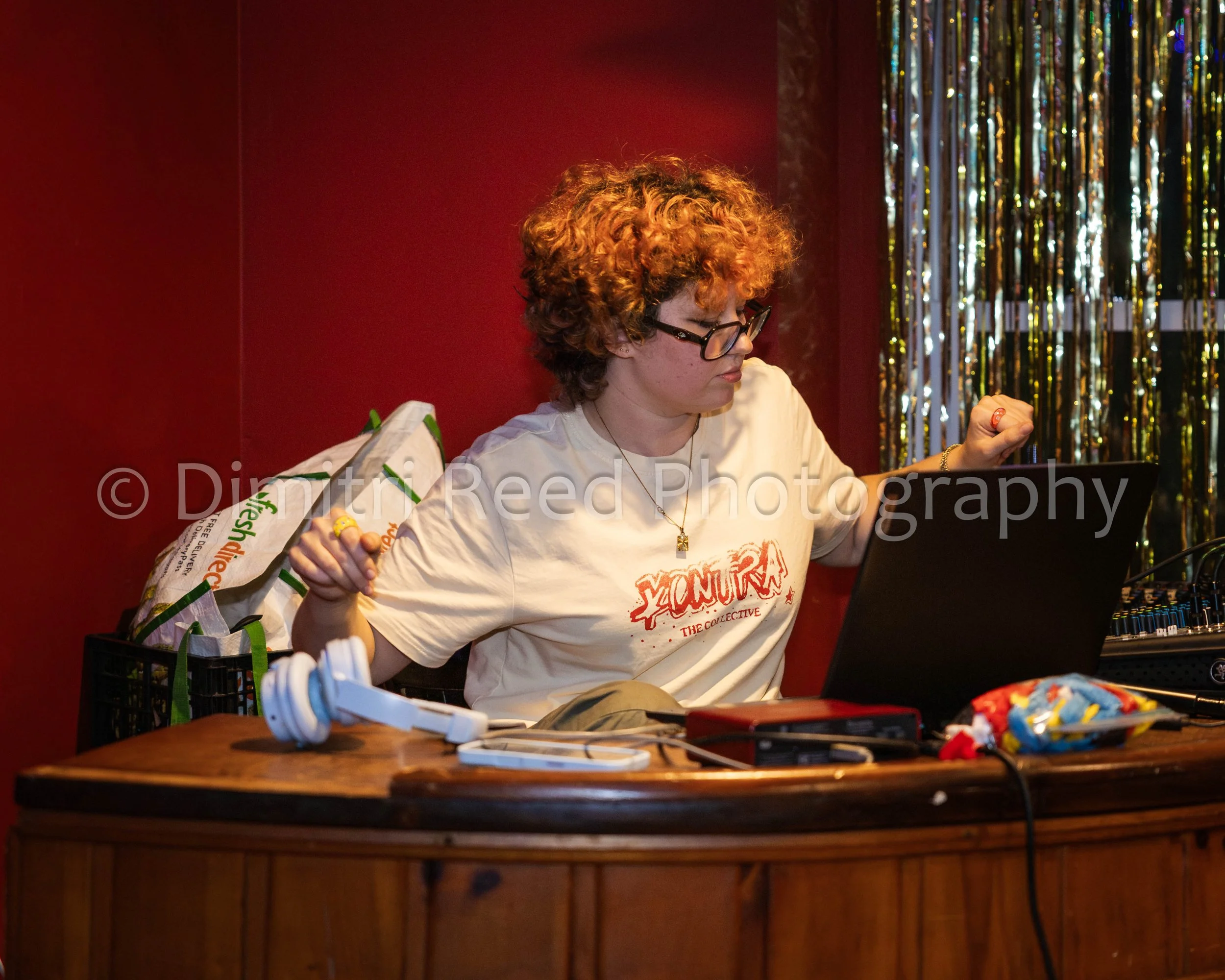 Person with curly red hair and glasses working at a desk with various items including headphones, a computer, and a bag, against a dark red wall with gold and silver streamers.